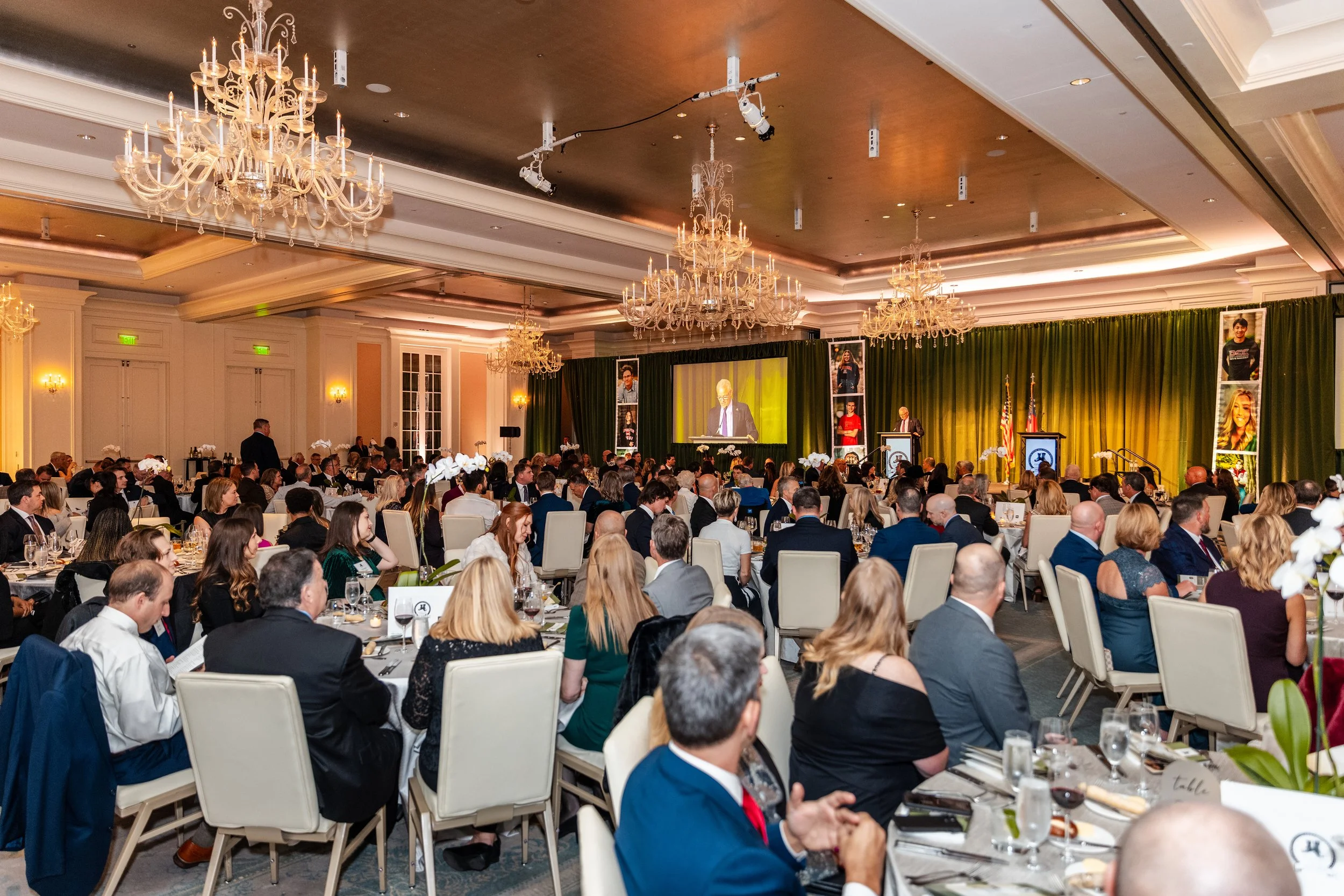 Large banquet hall filled with seated guests attending a formal event or conference, with a stage at the front, large screen, and decorated with chandeliers and floral arrangements.