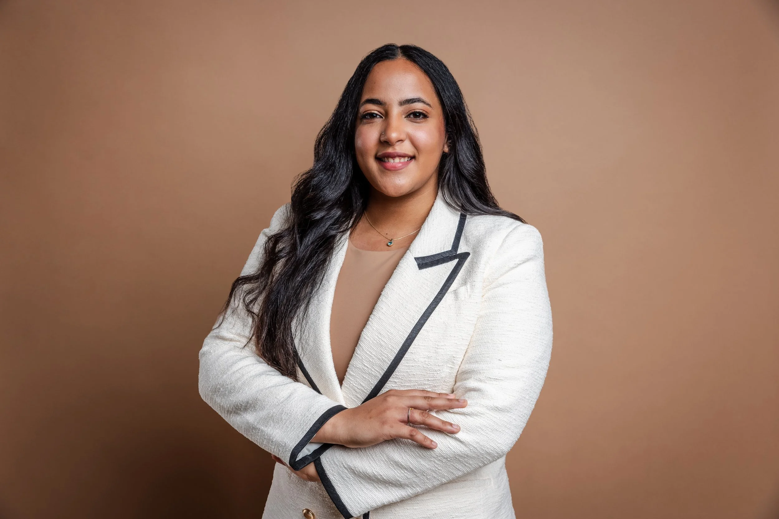 A smiling woman with long black hair wearing a white blazer with black trim, looking confident with her arms crossed, standing against a brown background.