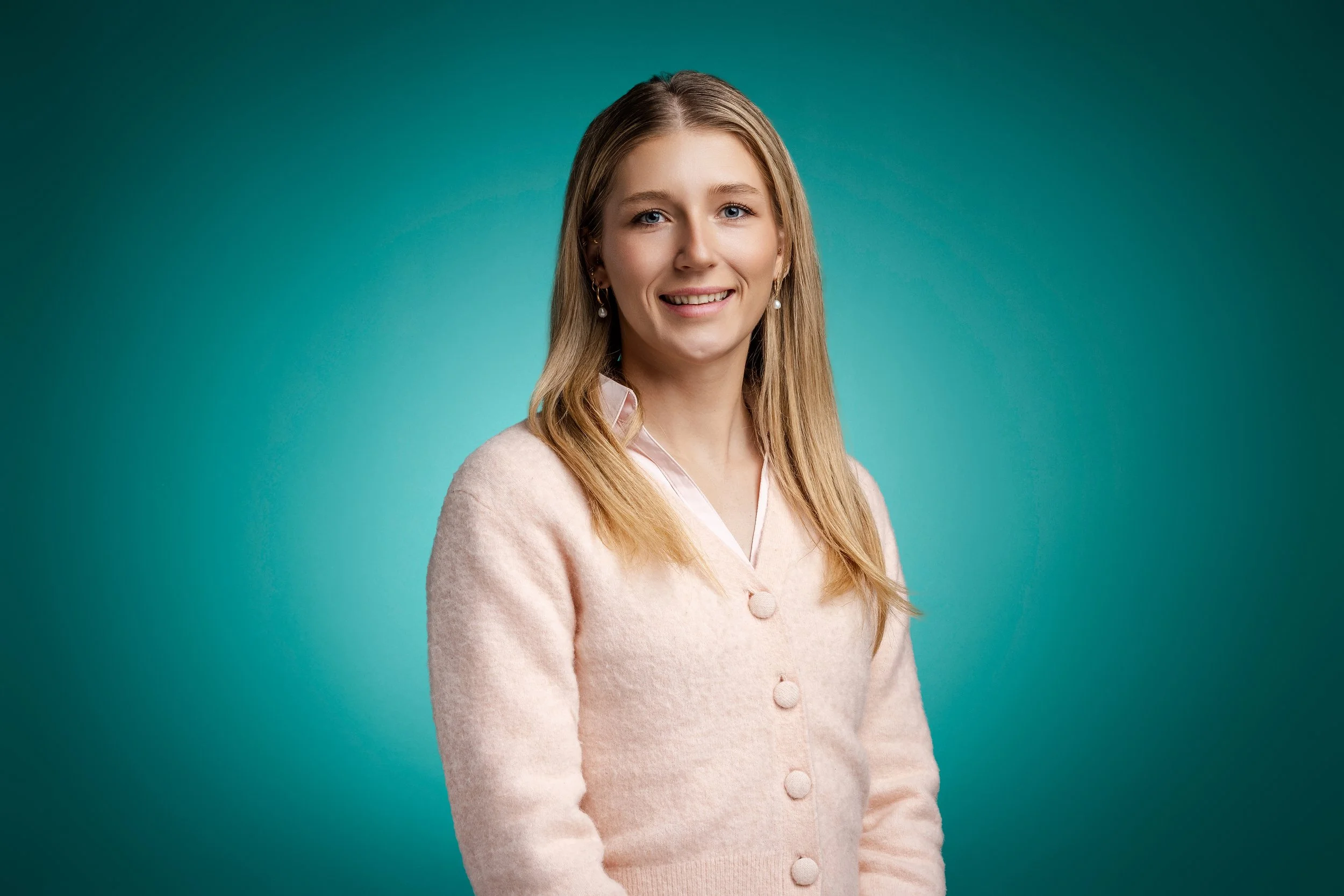 A young woman with blonde hair, wearing a light pink cardigan and pearl earrings, smiling in front of a teal background.