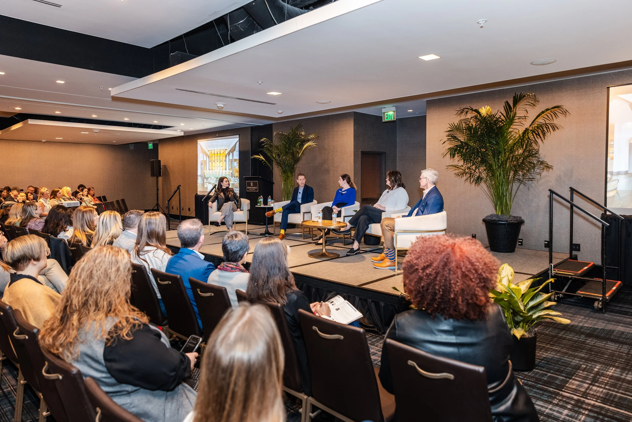 Panel discussion at a conference with five speakers on stage, audience members seated facing them, large potted plants, and a projection screen in the background.