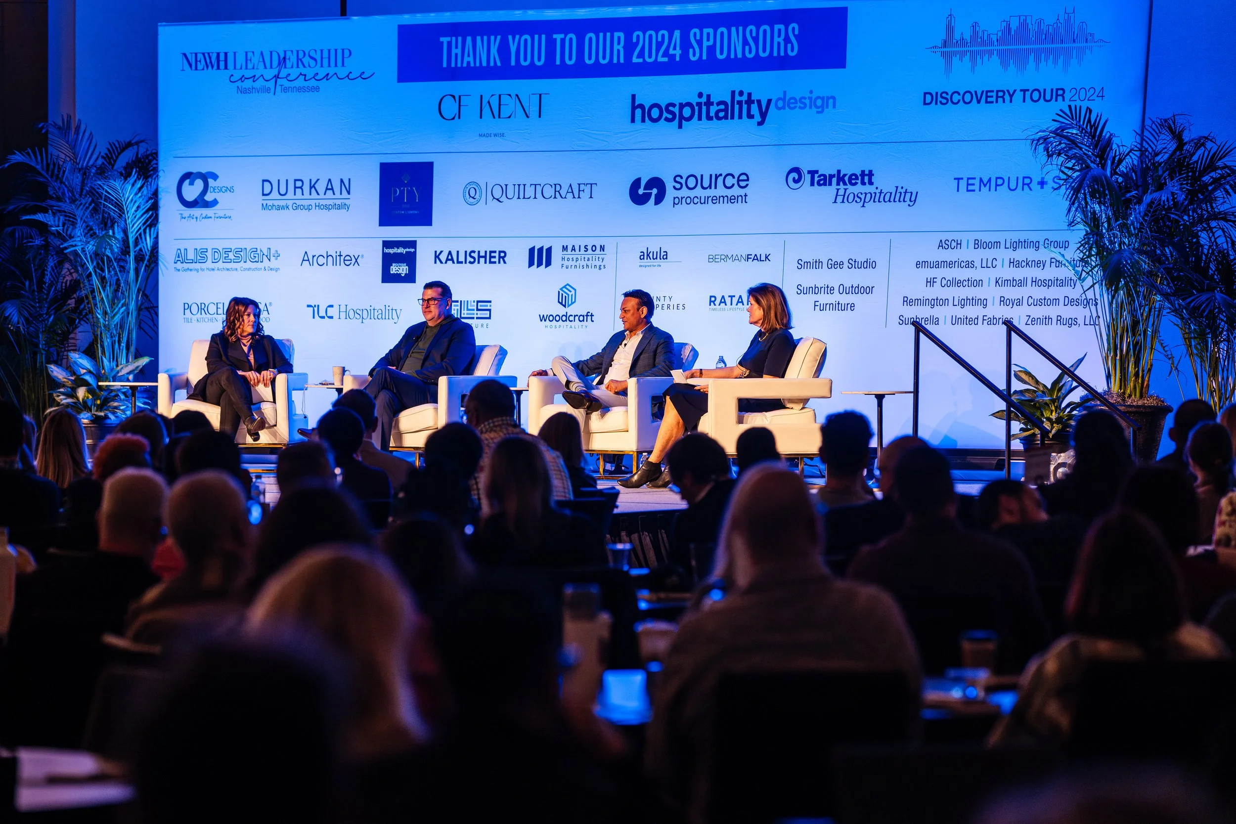 Panel discussion at a conference with four speakers on stage, four white armchairs, backdrop of sponsor logos, and audience members seated facing the stage.