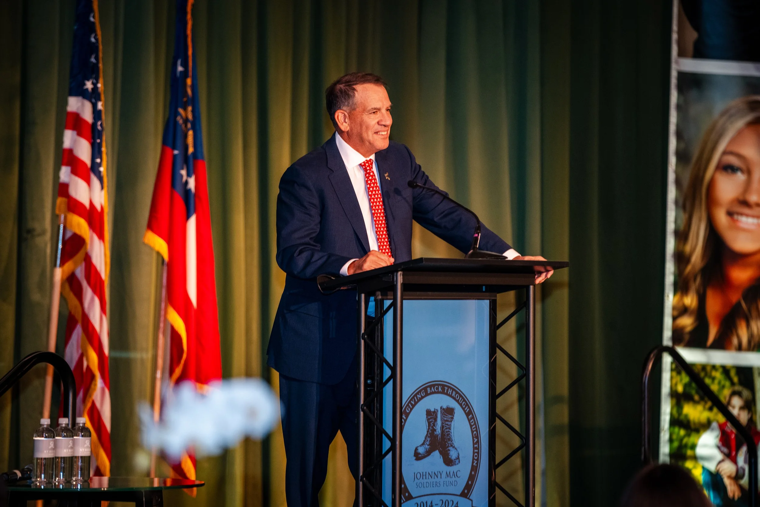 A man in a dark suit and red tie speaking at a podium with an organization logo, behind green curtains and American flags.