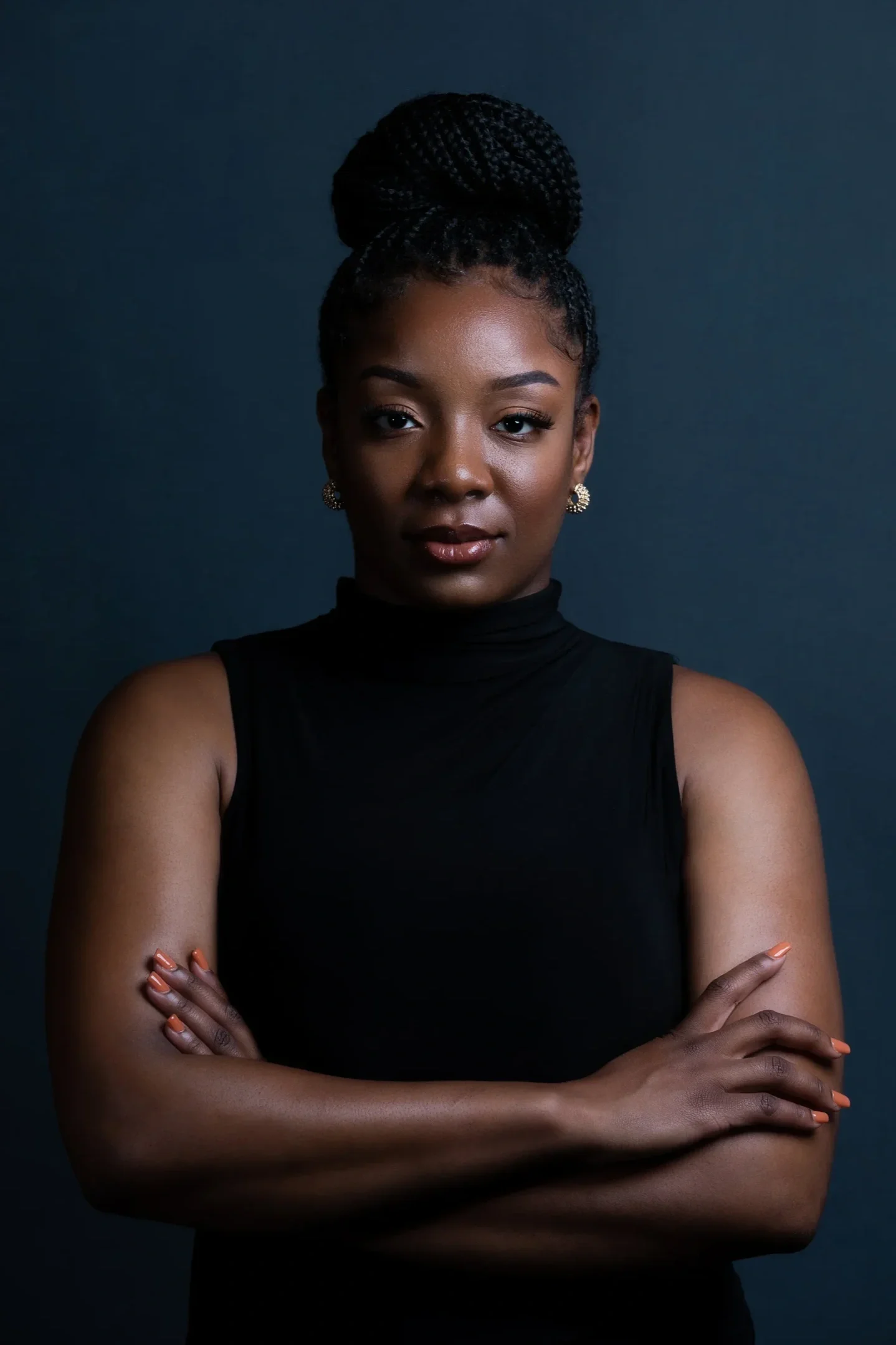 Portrait of a young Black woman with braided hair styled in a large bun, wearing a black sleeveless turtleneck, gold earrings, and light makeup, standing against a dark background.