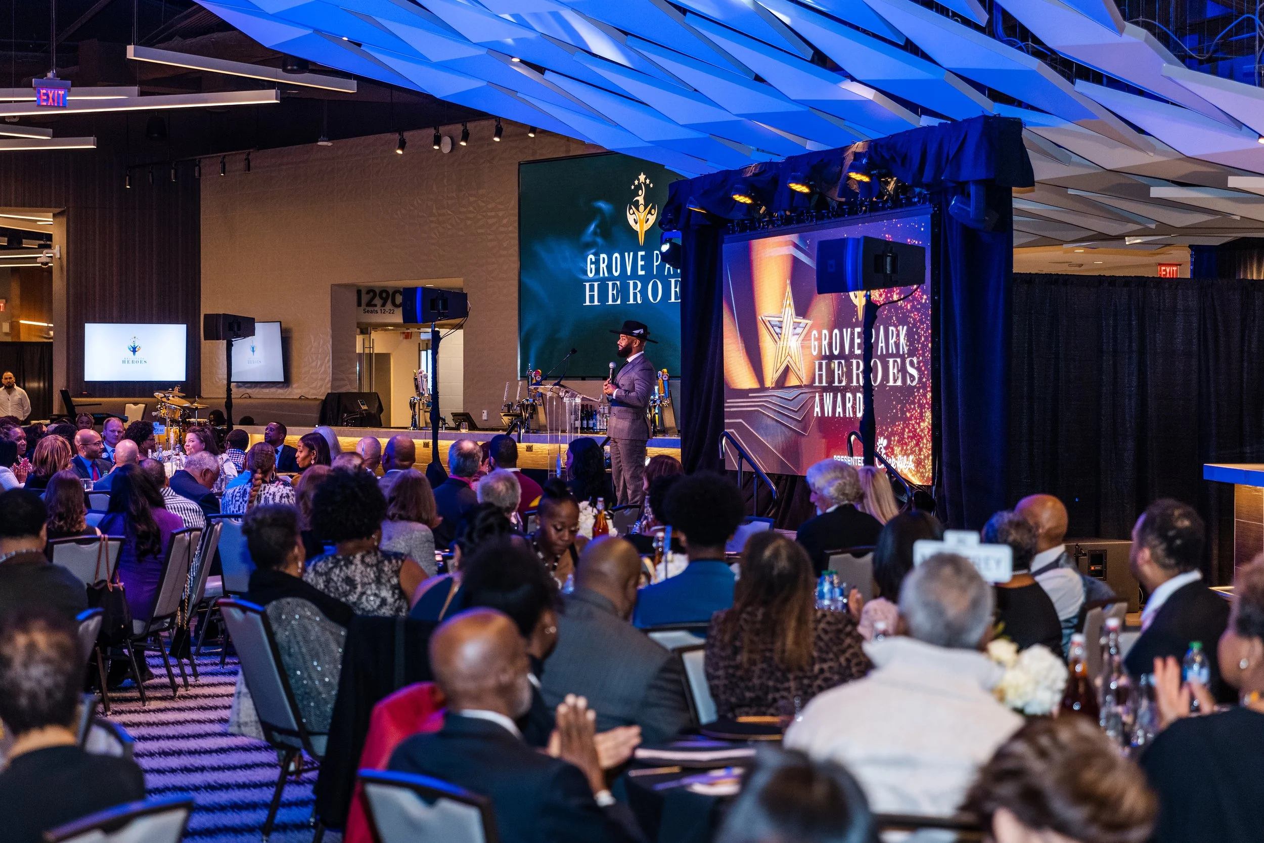 A large indoor event with a stage where a man in a gray suit and black hat speaks into a microphone. The stage has a large digital screen displaying the 'Grove Park Heroes Award' logo. The audience is seated at round tables, and there are multiple screens showing the same logo. The room has a modern interior with geometric ceiling panels and dim lighting.