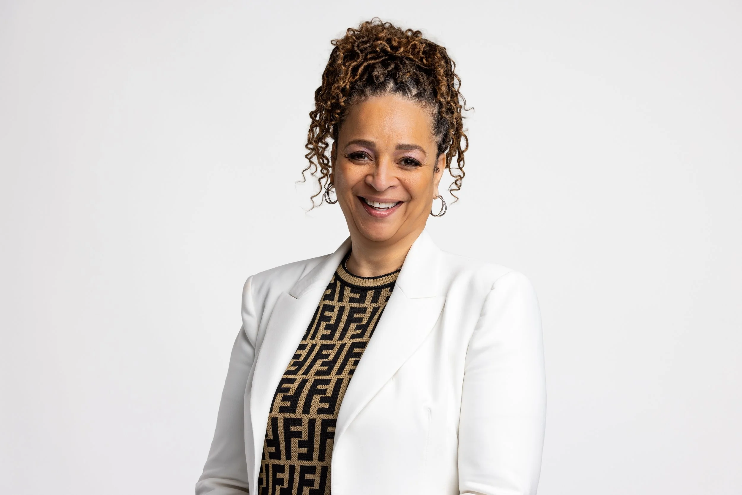 A woman with curly hair in a ponytail, wearing a white blazer over a patterned top, smiling against a plain white background.