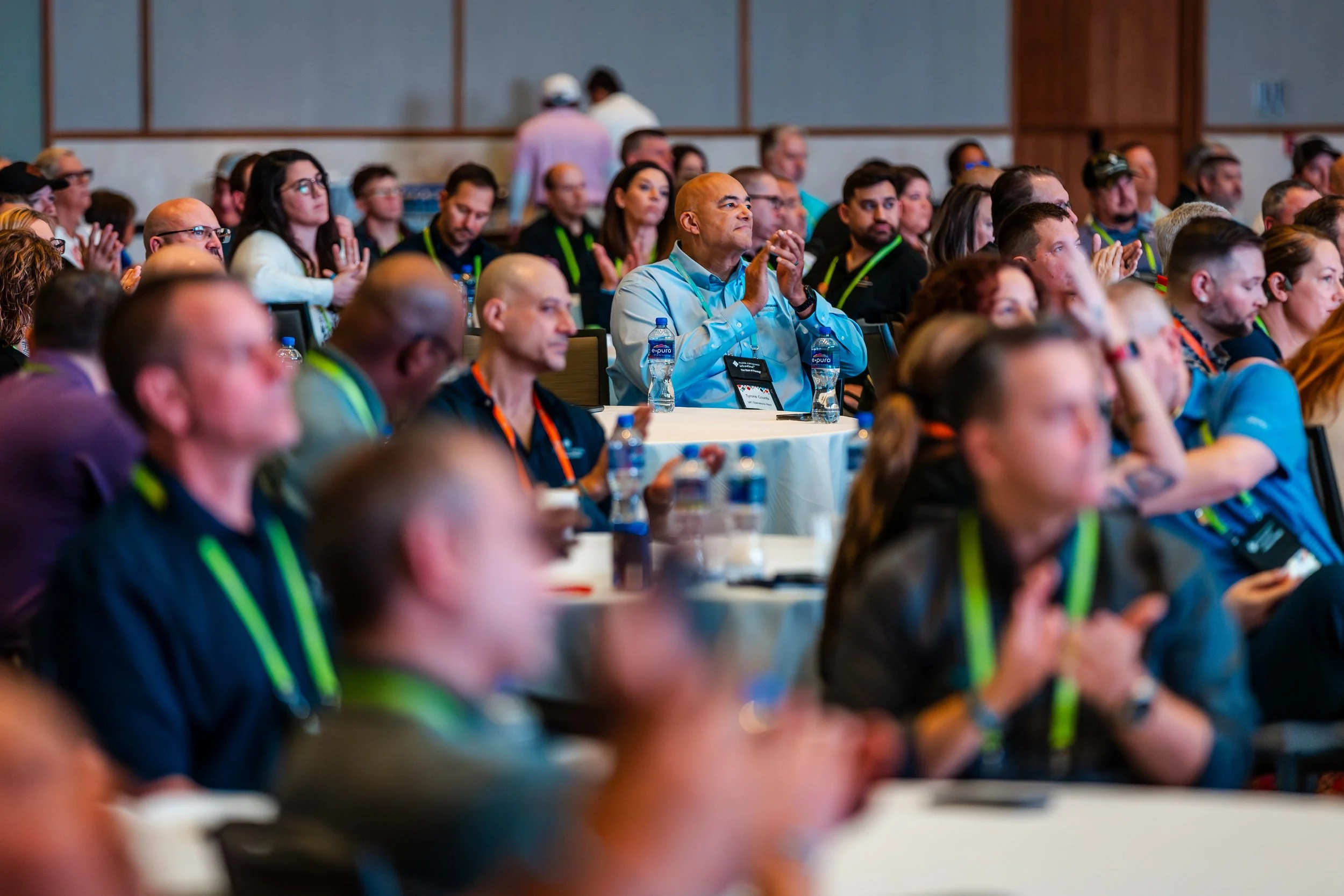 Attendees clapping and listening at a conference or seminar in a large room with many people of different backgrounds, some wearing conference badges and lanyards.