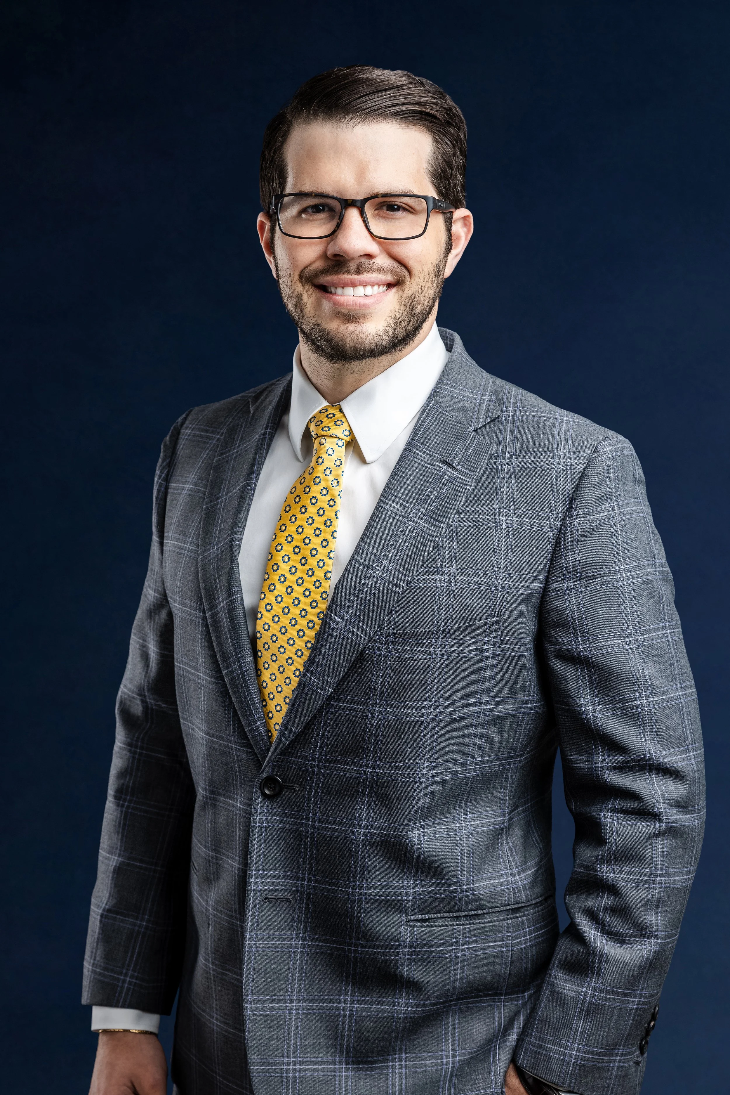 Portrait of a smiling man with dark hair and beard, wearing glasses, a gray plaid suit, white shirt, and yellow tie with a blue pattern, against a dark background.