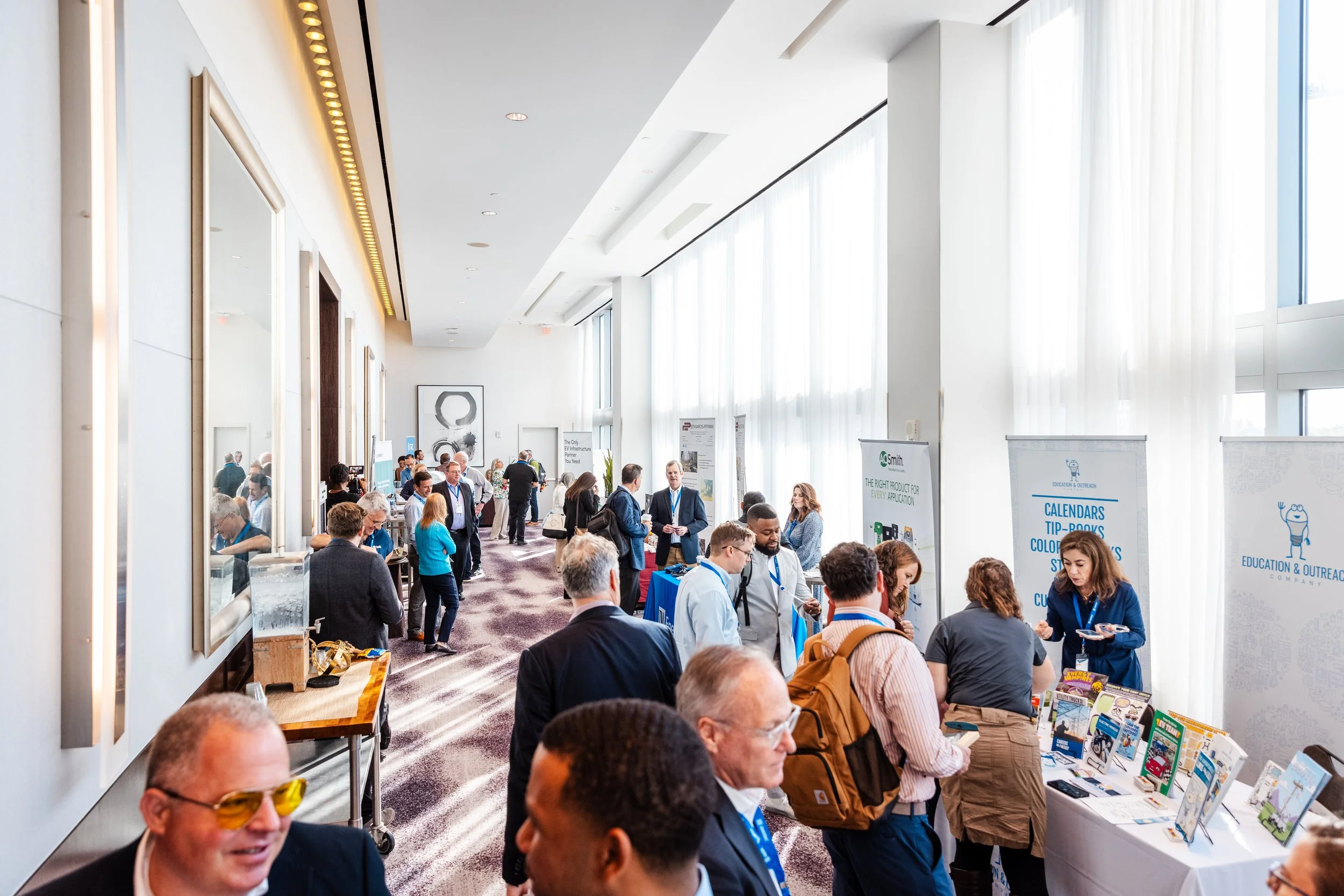 Conference or expo room with numerous attendees exploring booths and displays, large windows with white curtains allowing natural light.