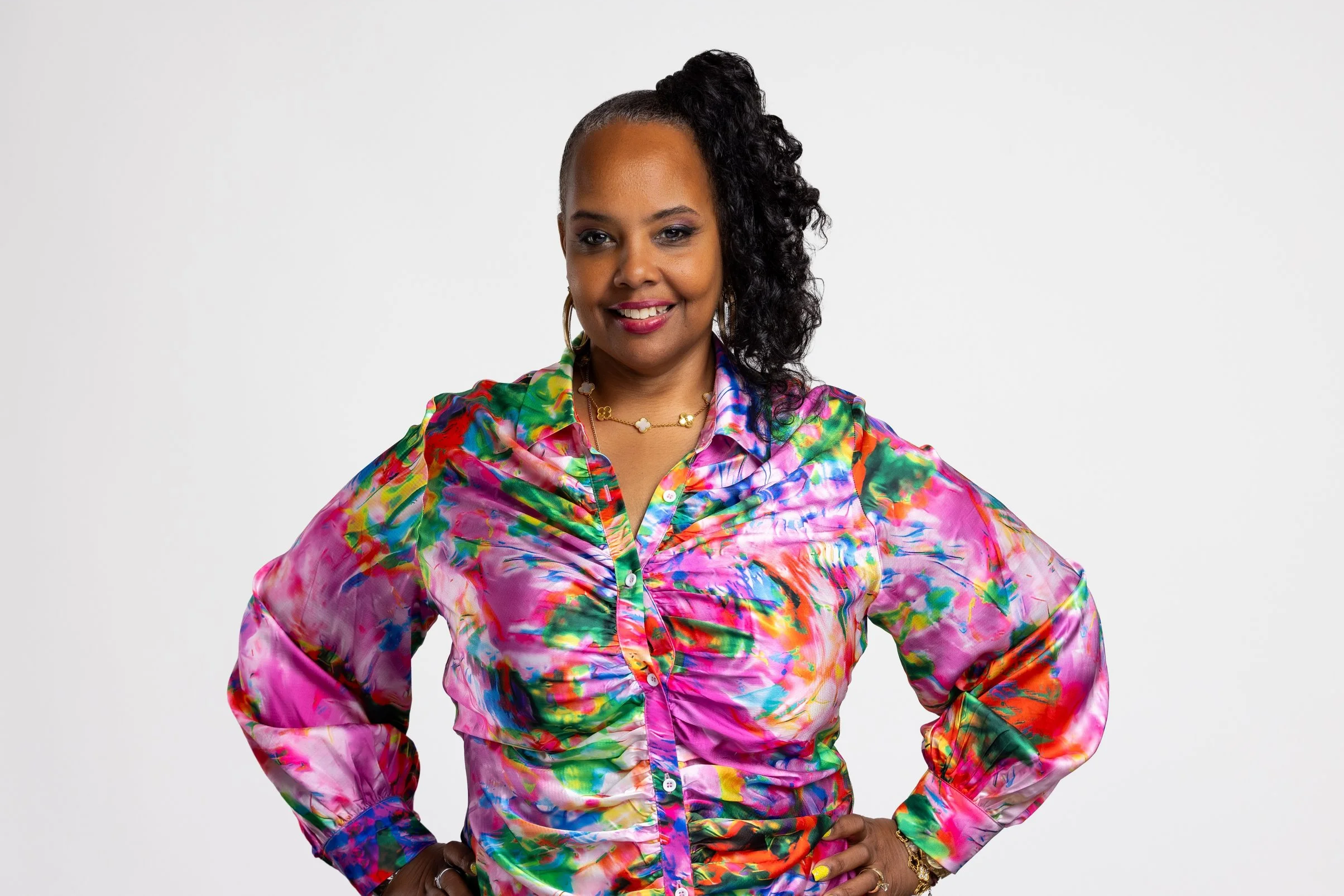 A woman with dark curly hair tied to one side, wearing a colorful, tie-dye style blouse and gold jewelry, standing confidently with her hands on her hips against a plain white background.