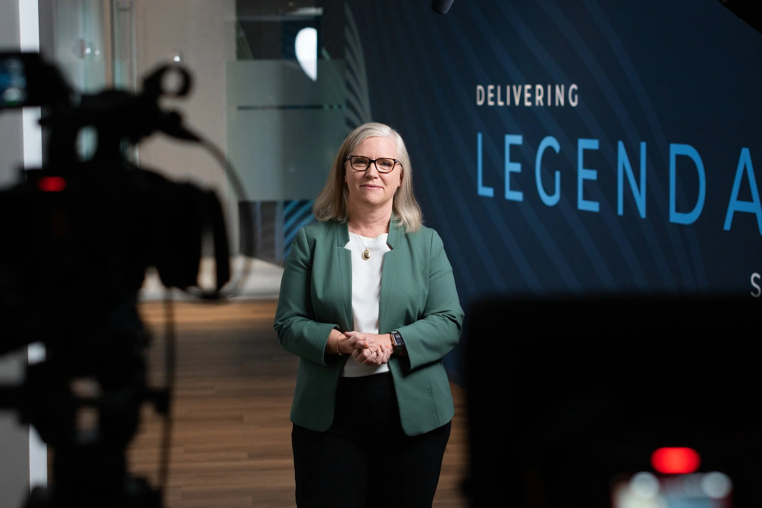 A woman with blonde hair, glasses, and a green blazer standing in front of a blue backdrop with the text 'Delivering Legendary' during a presentation or interview.