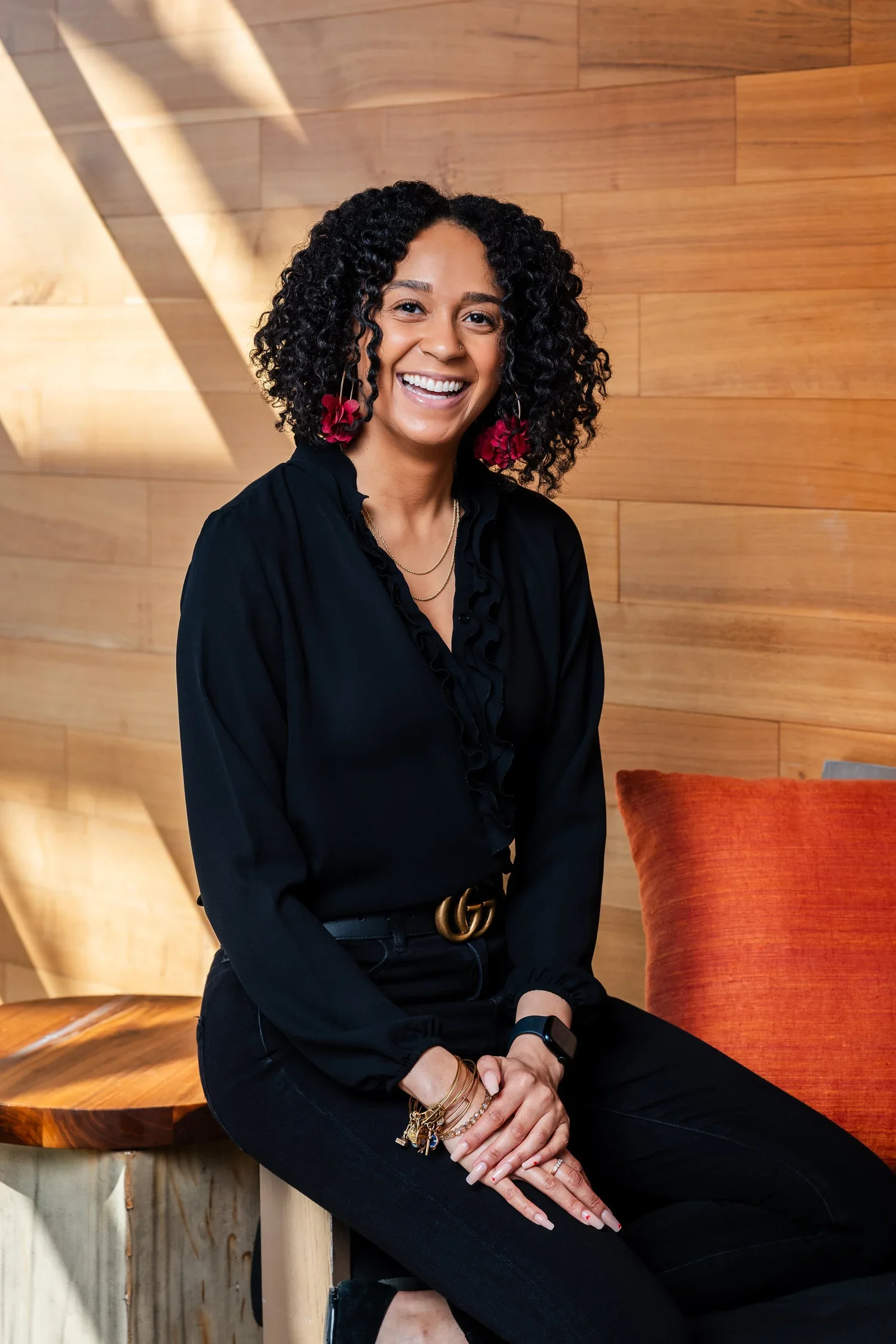 A smiling woman with curly black hair and red earrings, wearing a black blouse and black pants, sits on a wooden bench in front of a wood-paneled wall.