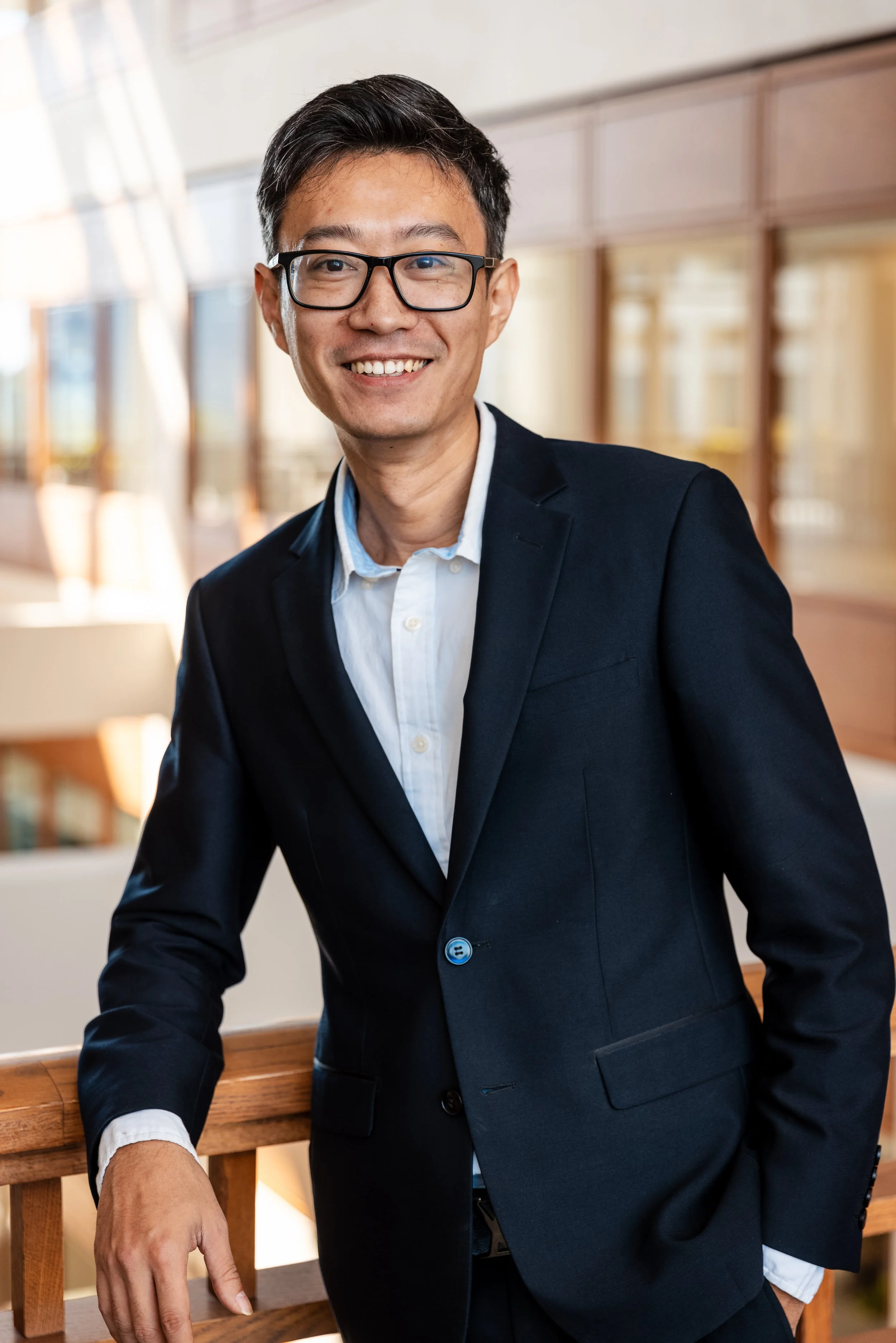 A smiling man with black hair and glasses, dressed in a black suit and white collared shirt, standing indoors with a blurred background of windows and wooden panels.