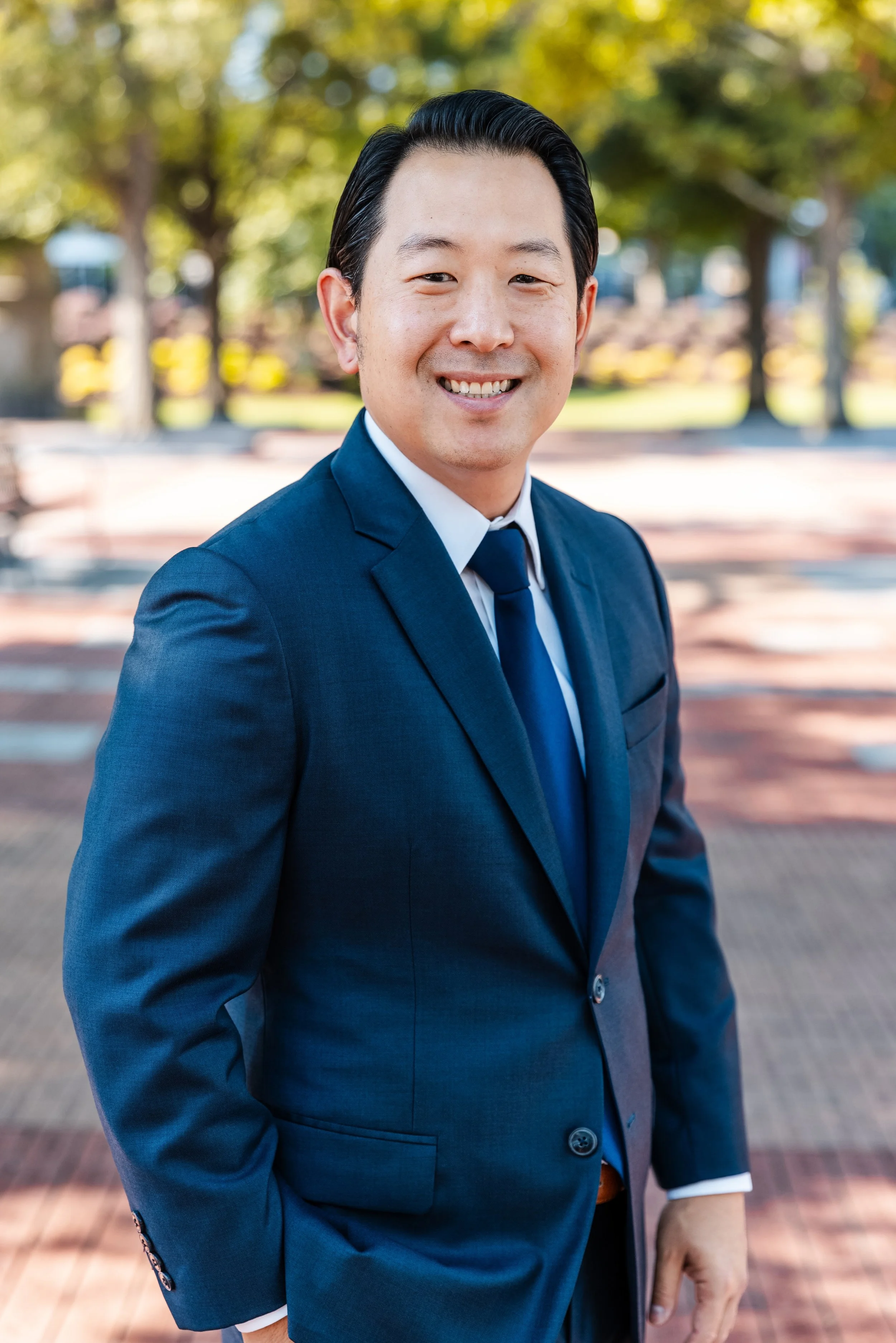 Smiling man in a blue suit standing outdoors in a park with trees and benches in the background.