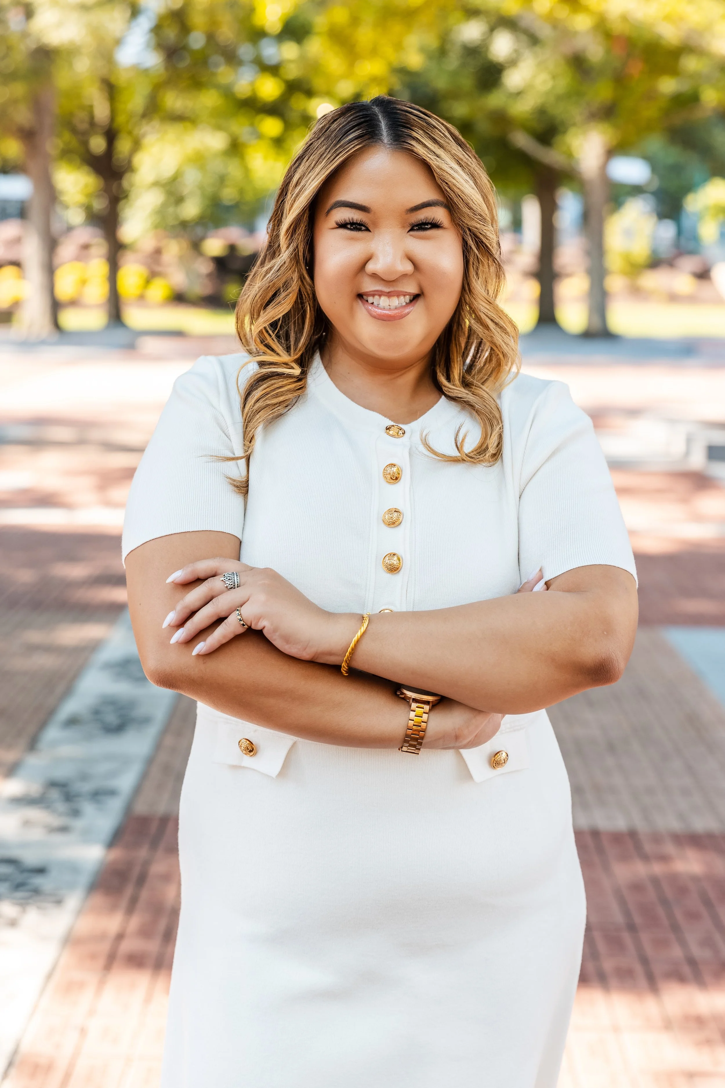 Smiling woman with wavy hair in a white dress standing outdoors with trees in the background.