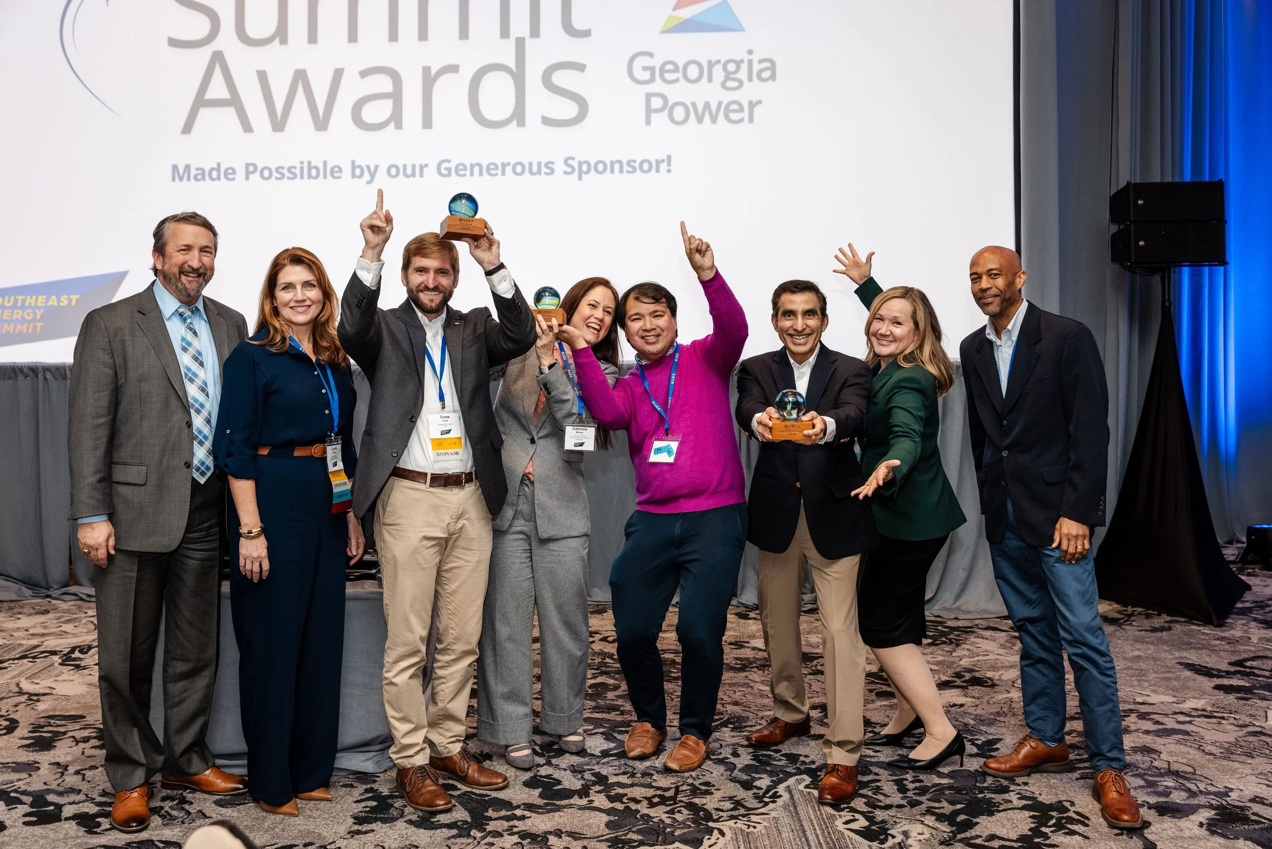 Group of eight people at an awards ceremony, some holding trophies, standing on stage in front of a large screen that reads 'Summit Awards Georgia Power,' smiling and celebrating.