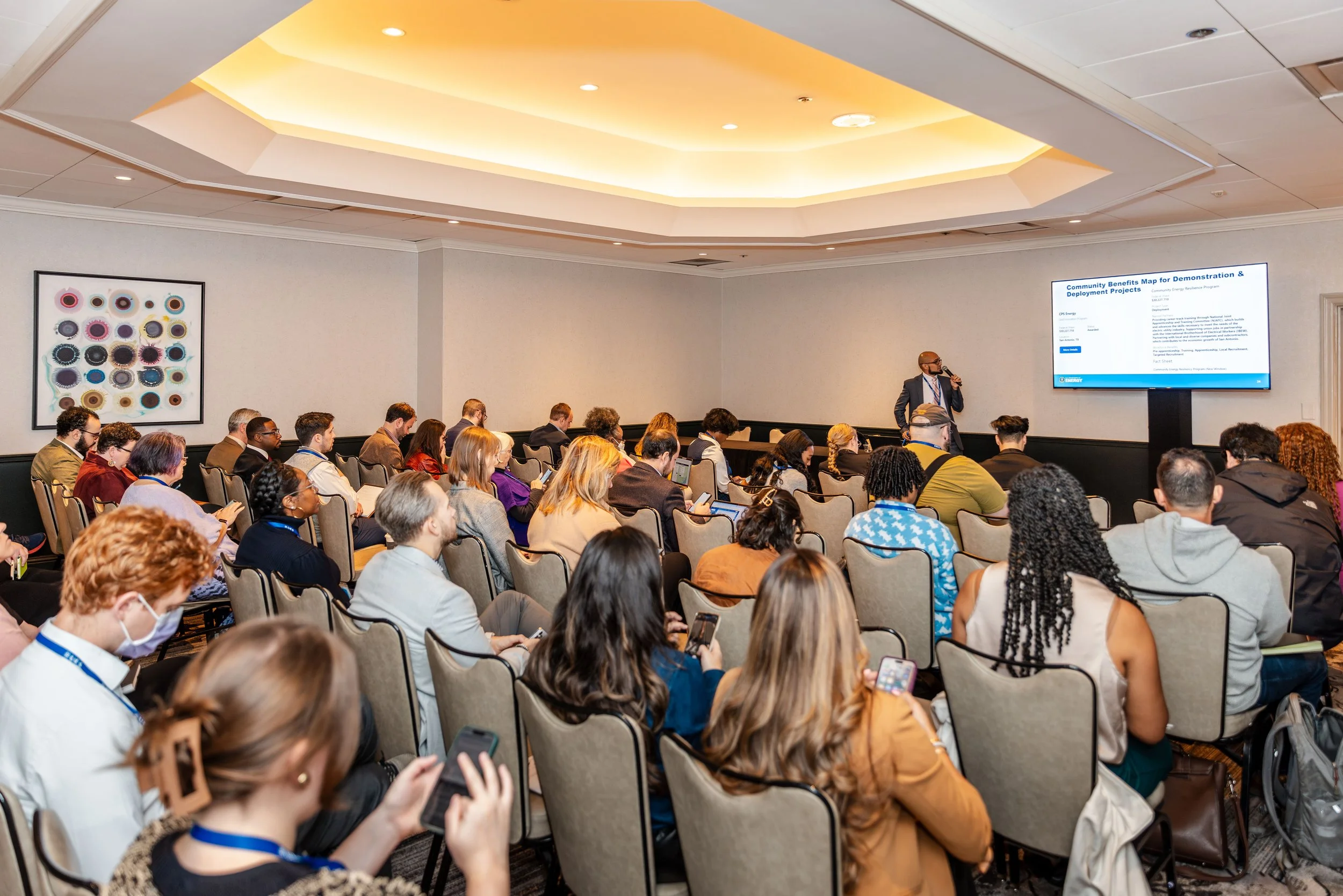 A conference room filled with a diverse group of people sitting in rows, attending a presentation. A speaker stands near a large screen displaying a presentation slide.