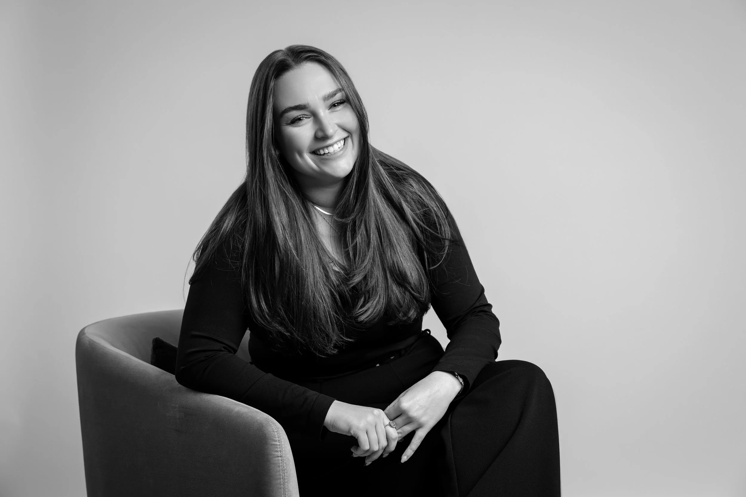 A smiling woman with long hair sitting on a chair in front of a plain background, in black and white.