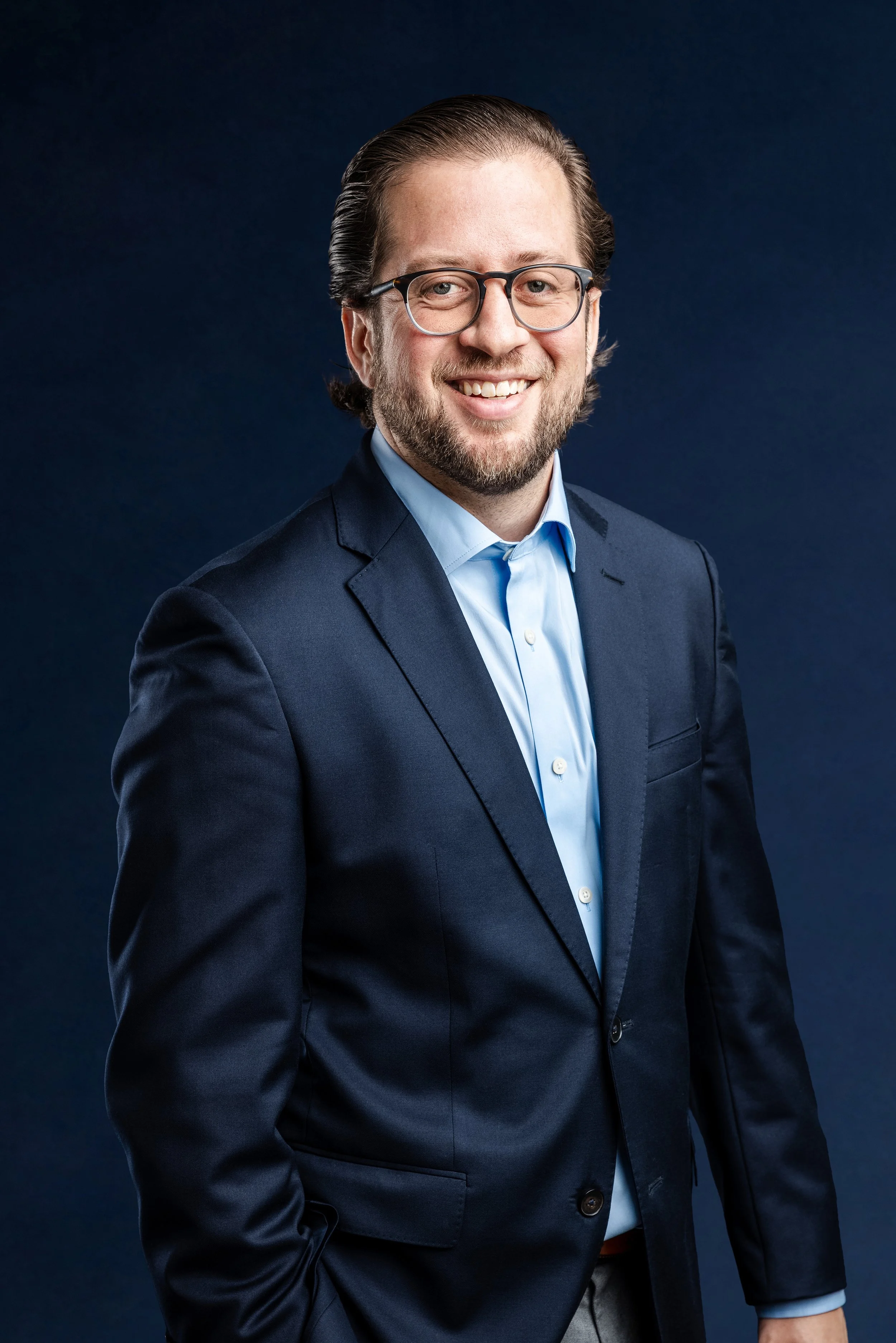A man with glasses, a beard, and brown hair smiling, dressed in a dark suit and light blue shirt, standing against a dark blue background.