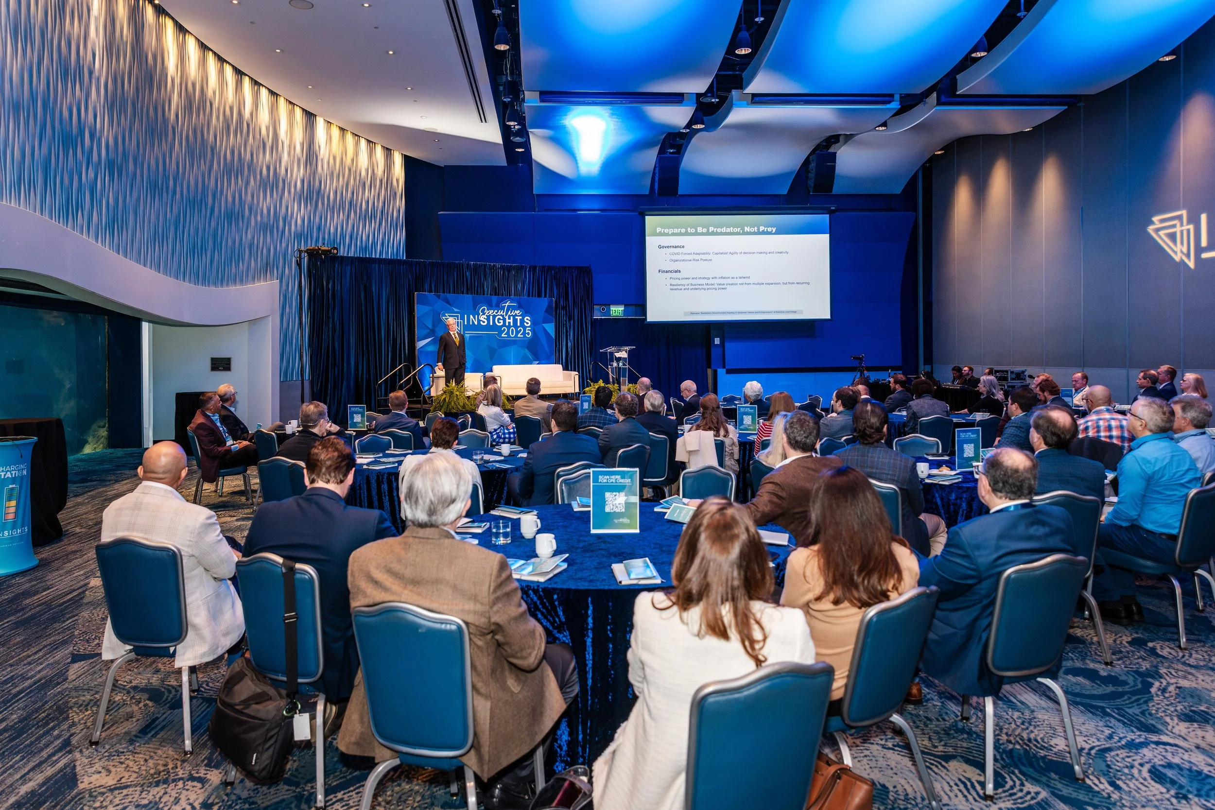 A conference or seminar in a large, modern auditorium with a speaker on stage and an audience seated at tables, listening and looking at presentation screens.