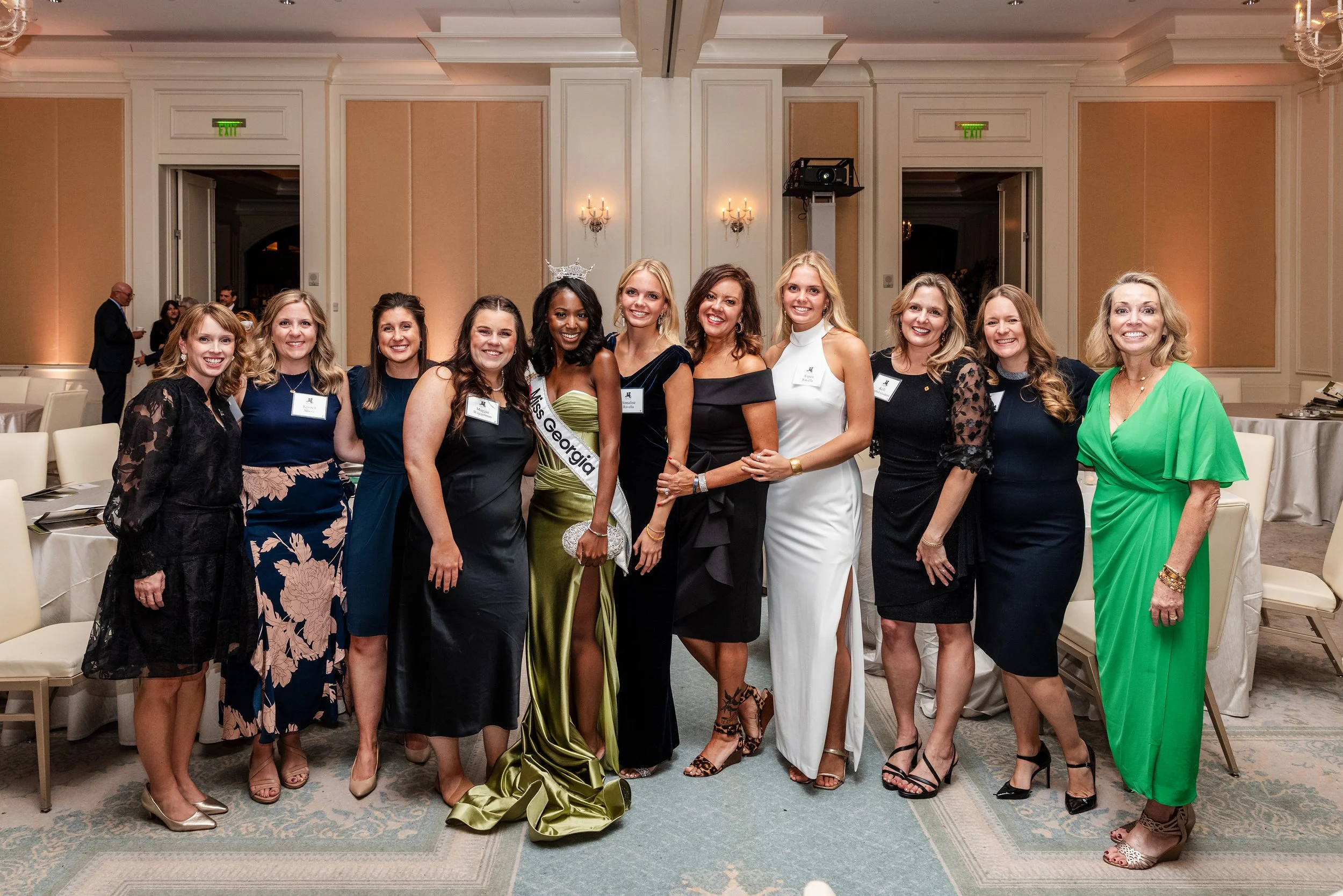 Group of women in formal attire at a social event in an elegant banquet hall, with one woman wearing a sash that reads 'Miss Georgia' and a crown, indicating a pageant or competition winner.
