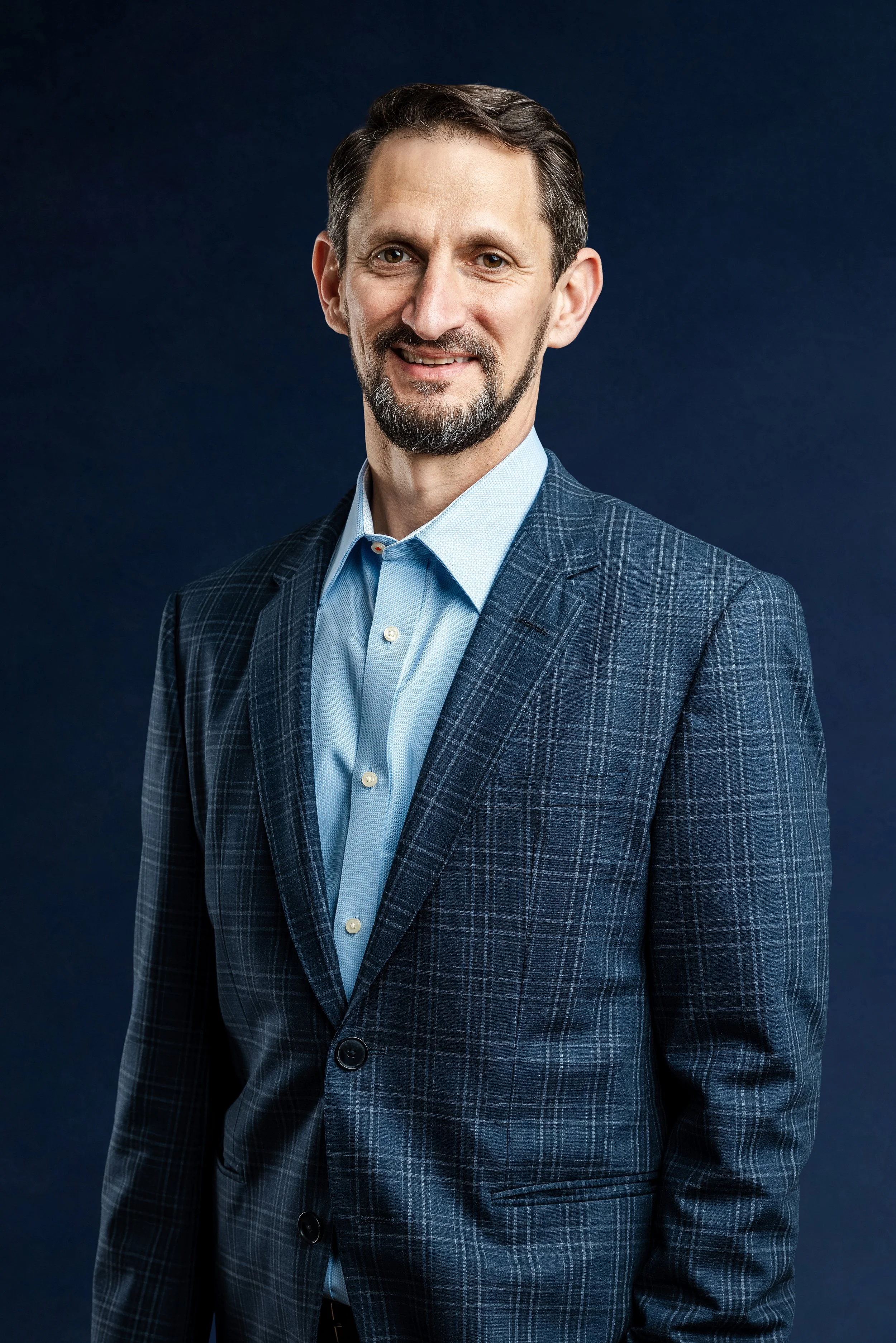 A middle-aged man with brown hair and a beard, wearing a blue checkered suit and light blue shirt, standing against a dark background and smiling at the camera.