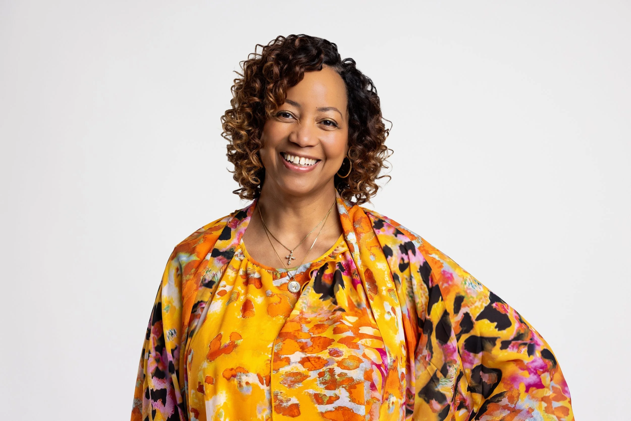 A smiling woman with curly brown hair wearing a colorful orange, pink, and black patterned top and a matching shawl, standing against a plain white background.