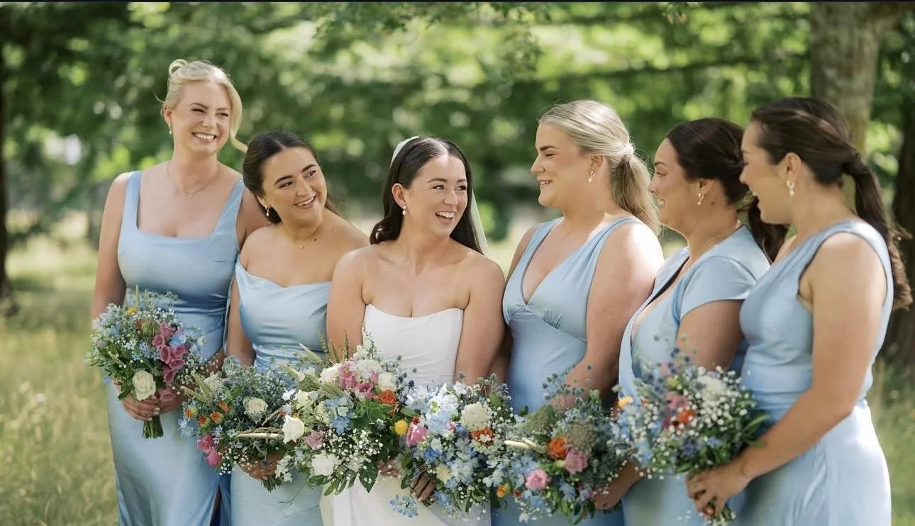 Group of women in blue dresses holding bouquets, standing outdoors in a park, smiling and talking.