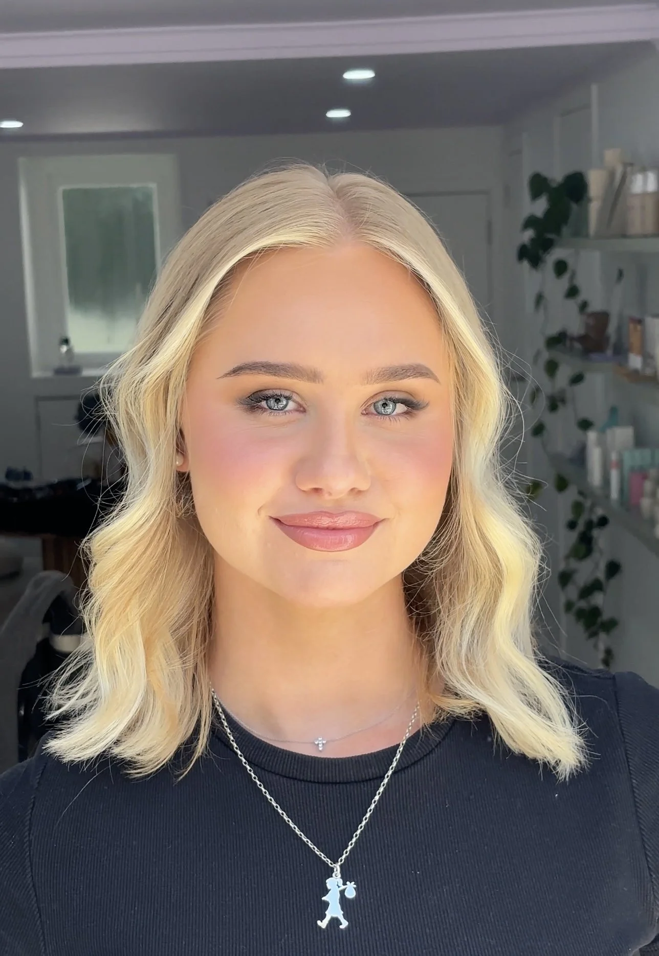 A young woman with blonde hair, blue eyes, and makeup, wearing a black shirt and two necklaces, smiling at the camera inside a room with shelves and a plant in the background.