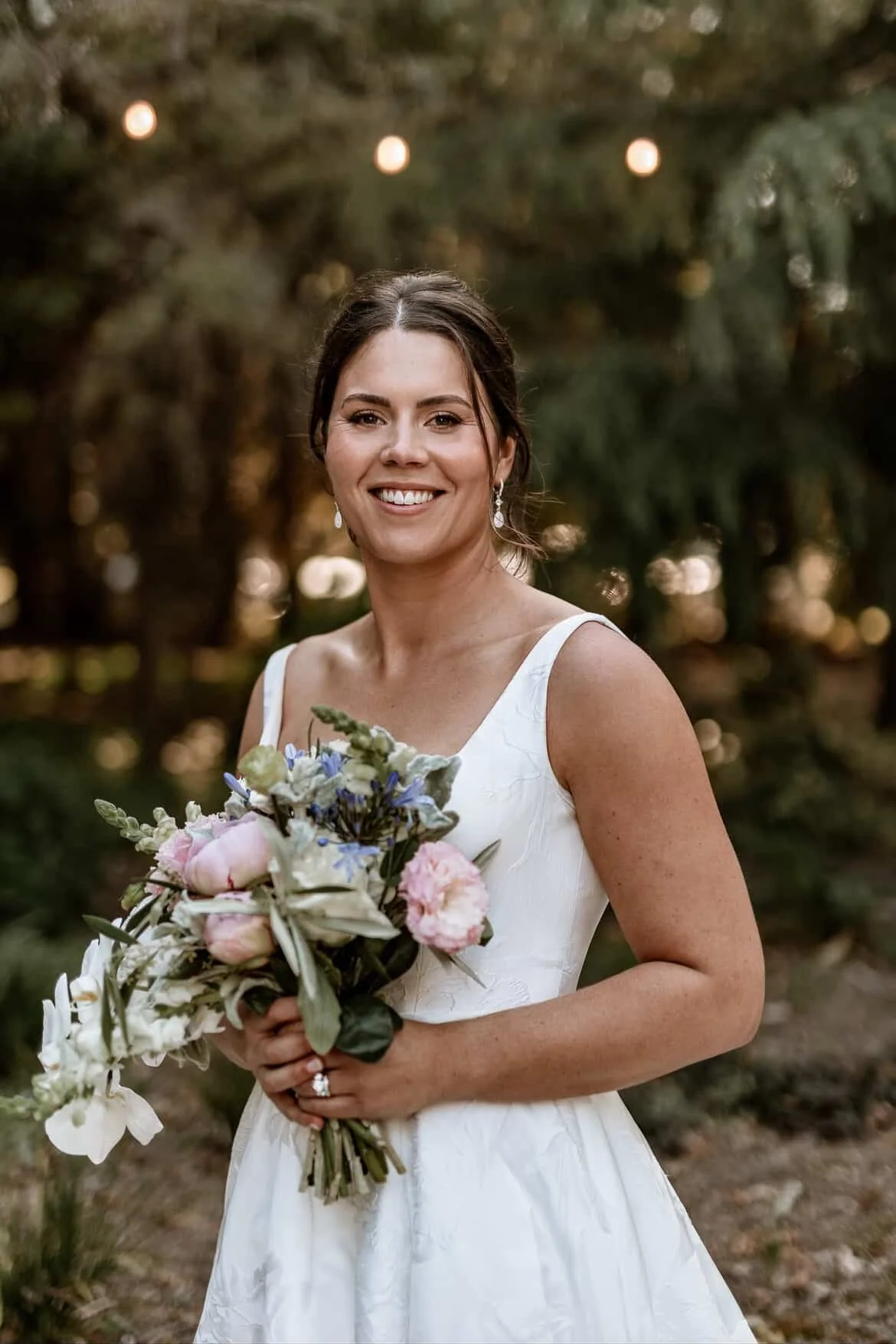 A smiling woman in a white wedding dress holding a bouquet of flowers outdoors with trees and string lights in the background.