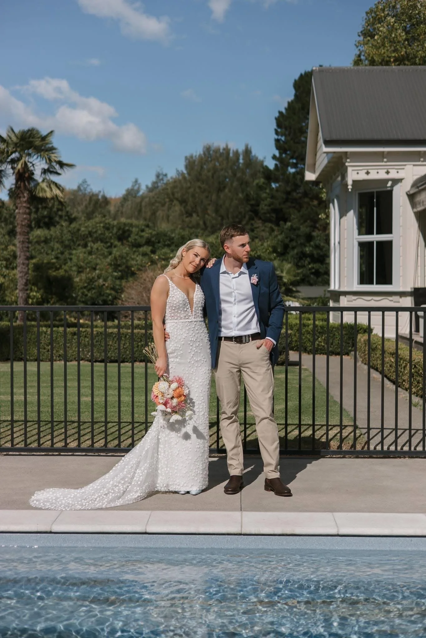 A bride and groom standing side by side outdoors near a swimming pool on a sunny day. The bride is holding a bouquet of pink and white flowers, and wearing a white wedding dress with textured details. The groom is dressed in a blue blazer, white shirt, and beige pants. They are next to a black metal fence, with green bushes and trees, and a white house with a dark roof in the background.