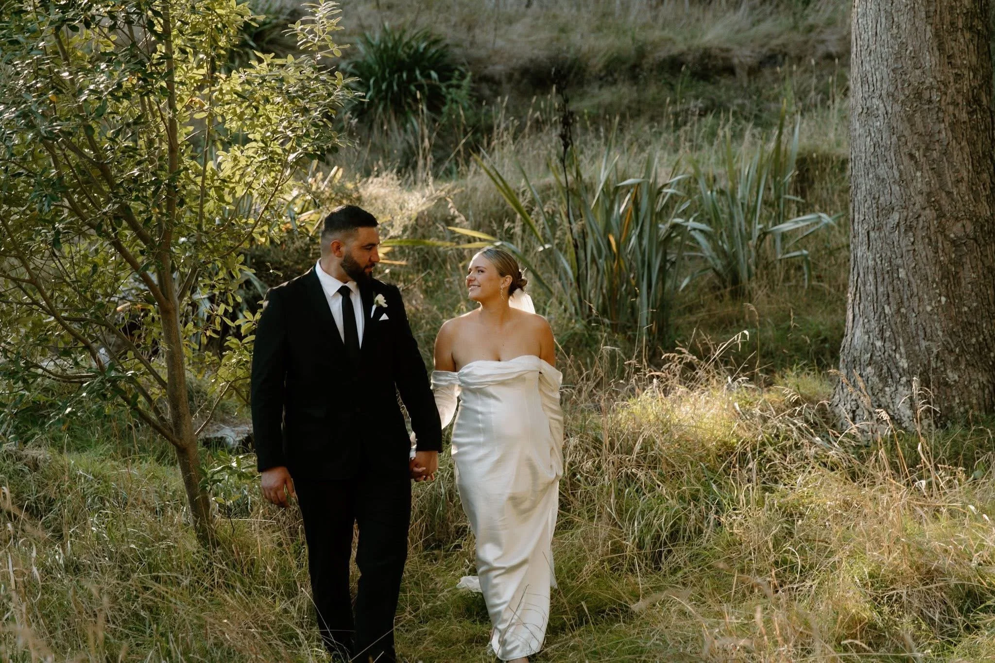 A newlywed couple walking hand in hand through a grassy, wooded area during daytime. The groom is wearing a black suit with a white shirt and black tie. The bride is wearing a white off-the-shoulder wedding gown. Both are smiling and looking at each 