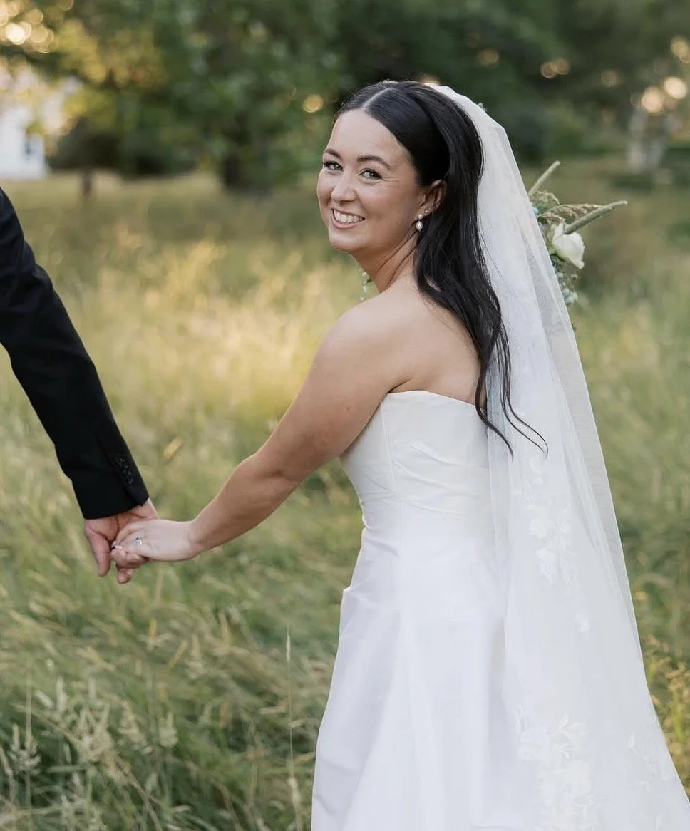 A smiling bride in a white strapless wedding dress and veil holding hands with her groom in a black suit outdoors.