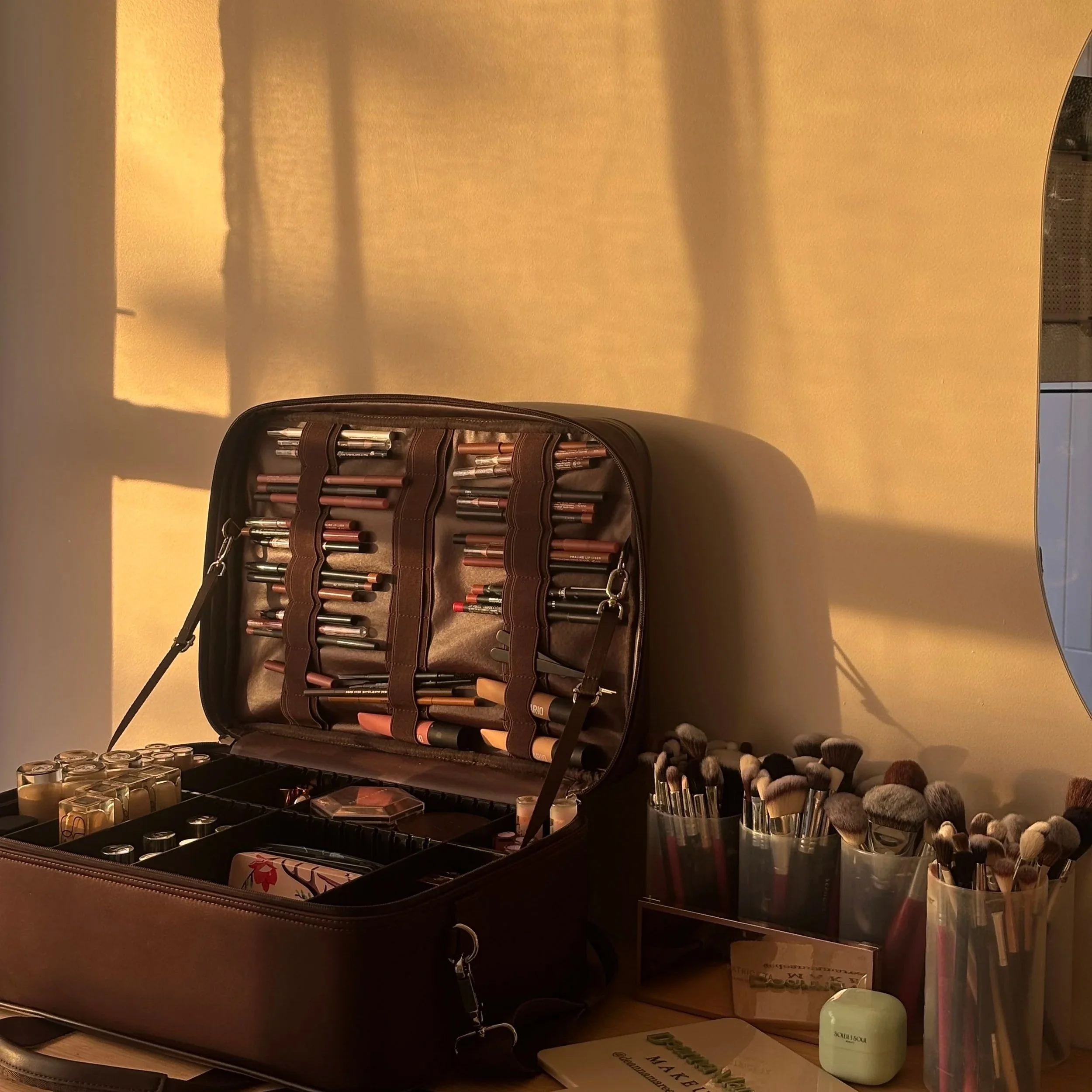 A brown makeup case with various brushes and makeup products, illuminated by warm sunlight, placed on a table with additional makeup brushes in containers nearby.