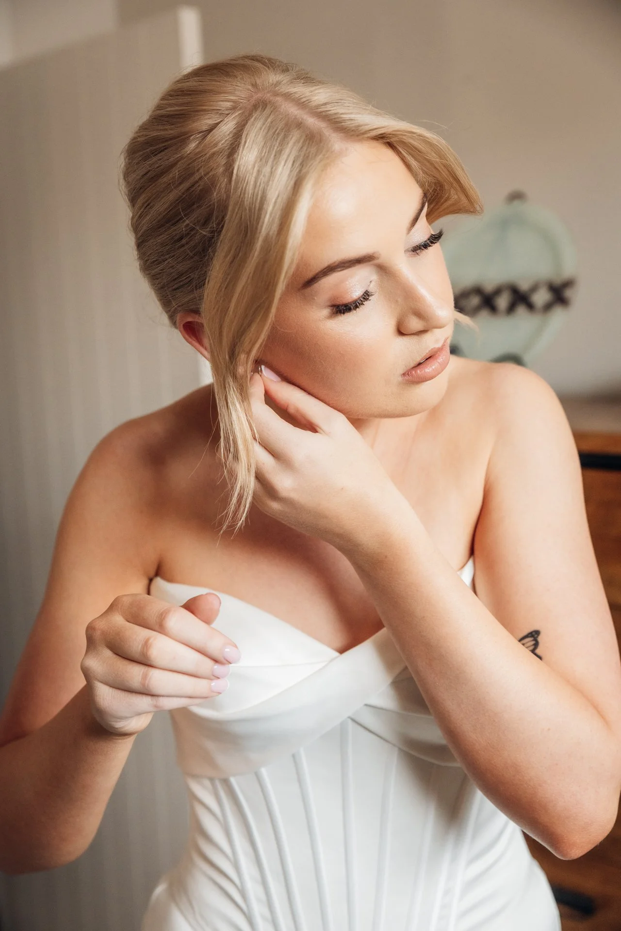 A woman with blonde hair styled in an updo, wearing a strapless white dress, is touching her hair and looking down with closed eyes.