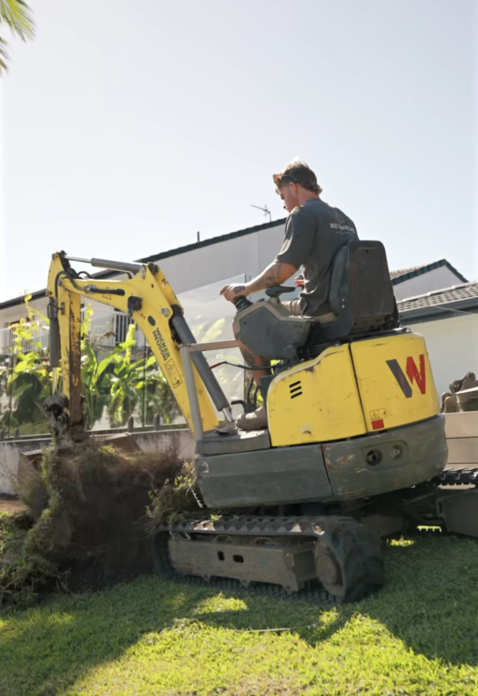 A person operating a small yellow excavator digging in a garden, with a house and plants in the background.