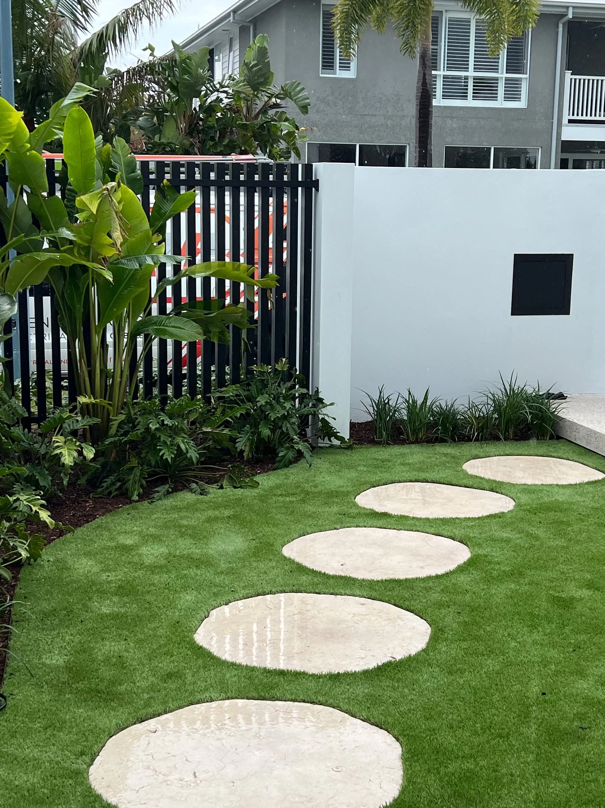 A modern backyard with a manicured lawn, circular stepping stones, lush green plants, and a white fence with a black lattice section.