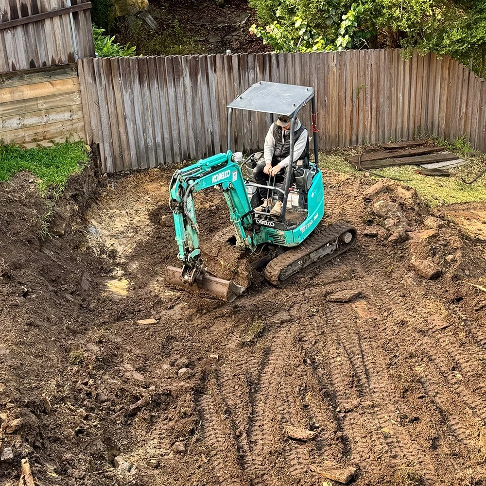 A person operating a small blue Komatsu mini excavator on a dirt construction site next to a wooden fence.