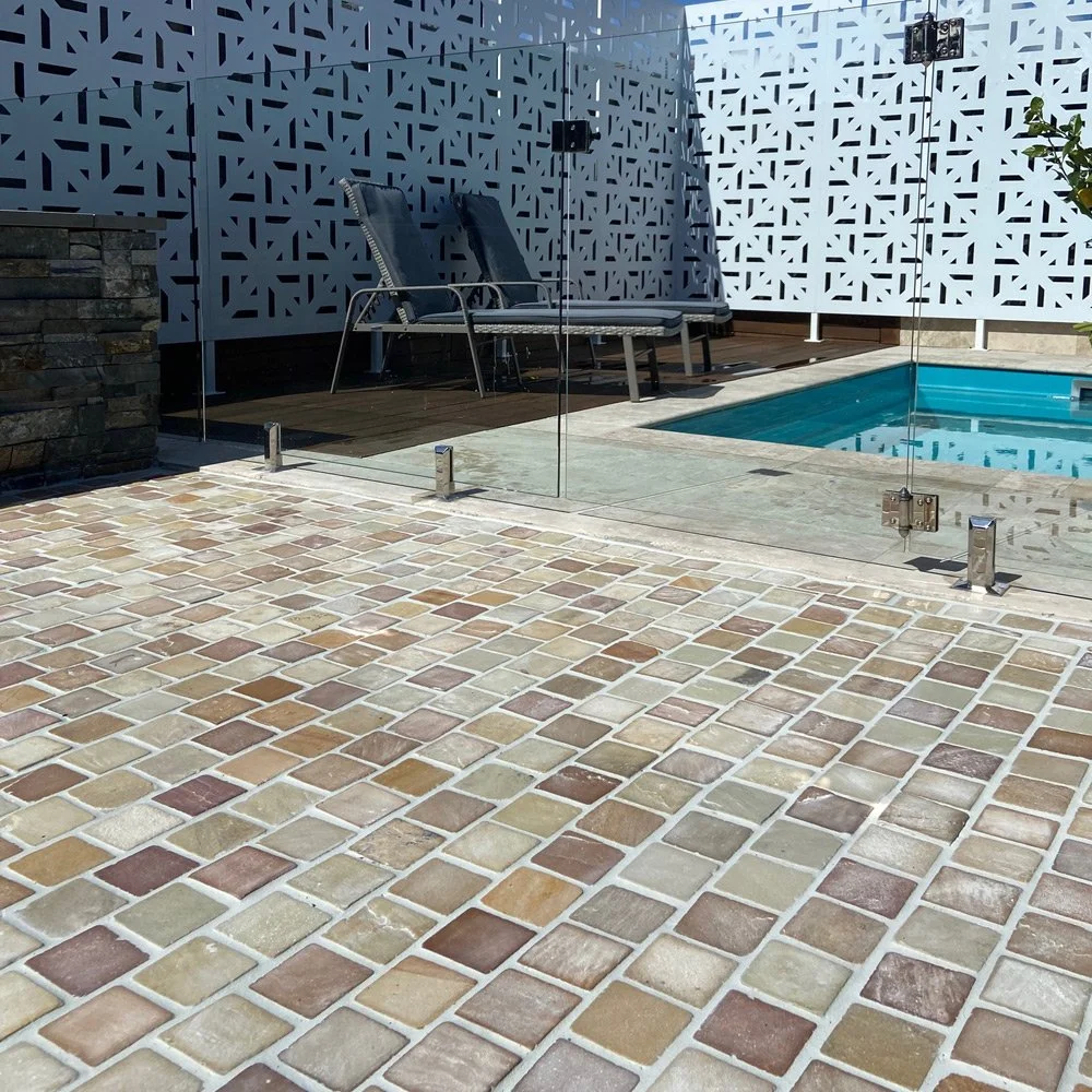 A small backyard pool enclosed by a glass fence, with two lounge chairs and a decorative white perforated privacy screen in the background, surrounded by brick-paved flooring.