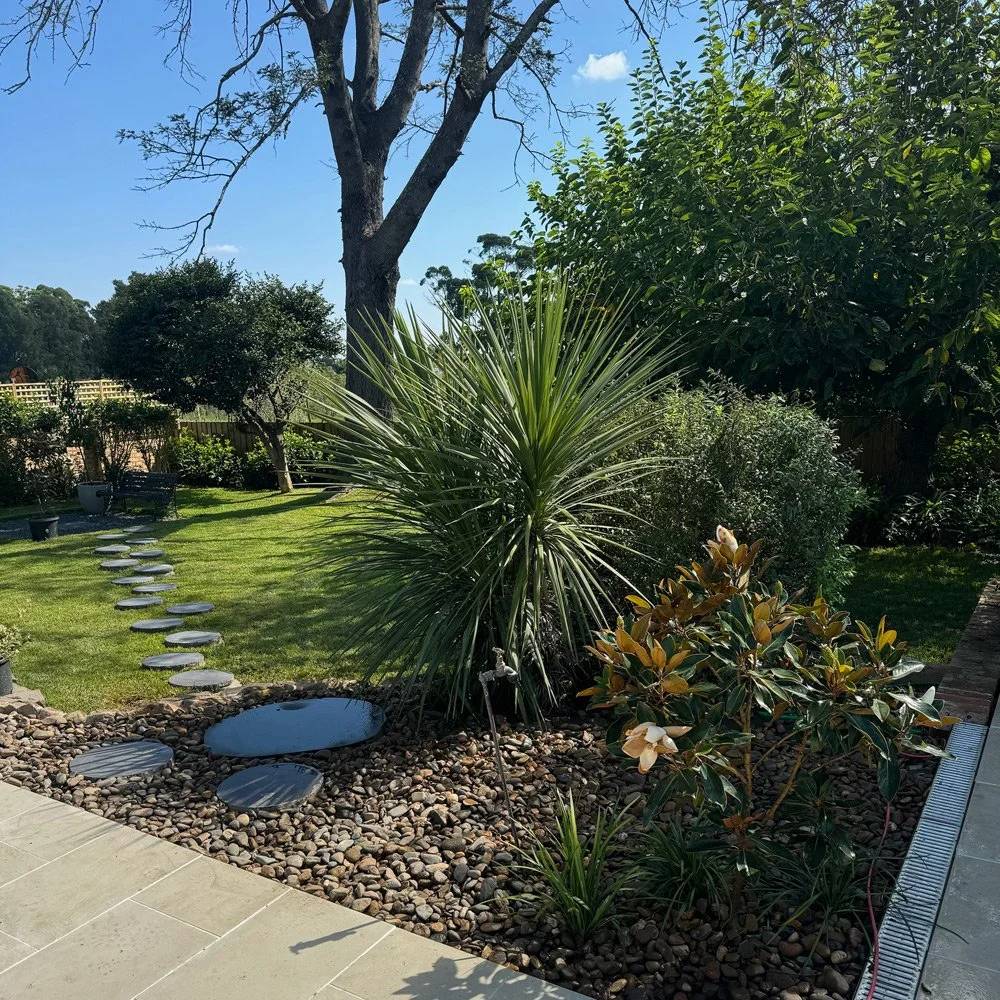 A backyard garden with a stepping stone path, trees, bushes, and a well-maintained lawn, under a clear blue sky.