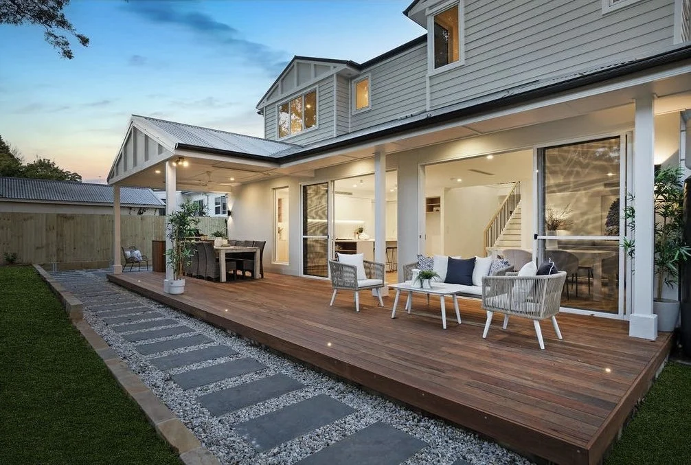 A backyard patio with outdoor furniture, including a dining table and a seating area, attached to a two-story house with large glass sliding doors, surrounded by a wooden fence and a walkway made of stone pavers.
