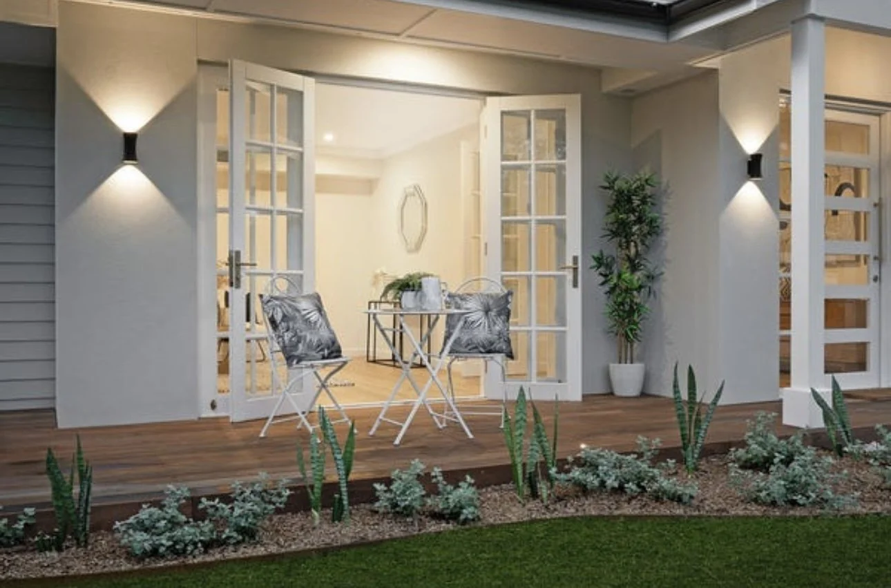 A cozy outdoor patio with a small table and two chairs, with open glass doors, surrounded by a garden with green plants and lighting at night.