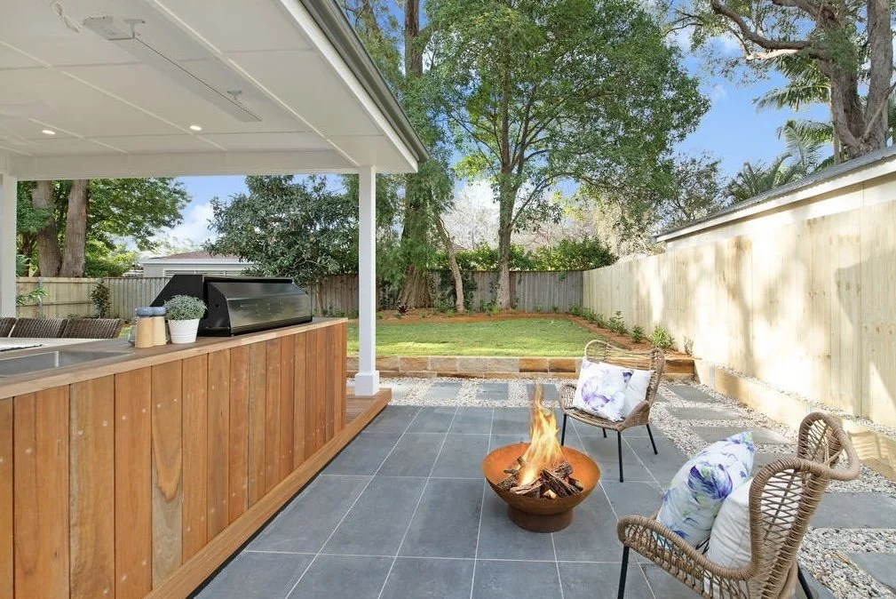 Outdoor patio with a fire pit, two chairs with cushions, a wooden bar, and a lawn with trees and a fence in the background.