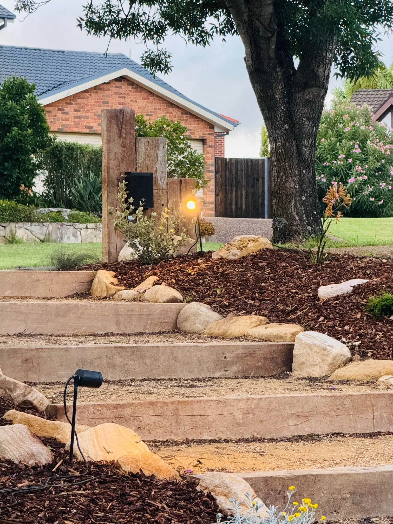 Outdoor garden with stone steps, rocks, plants, a large tree, a wooden fence, a brick house, and outdoor lighting.
