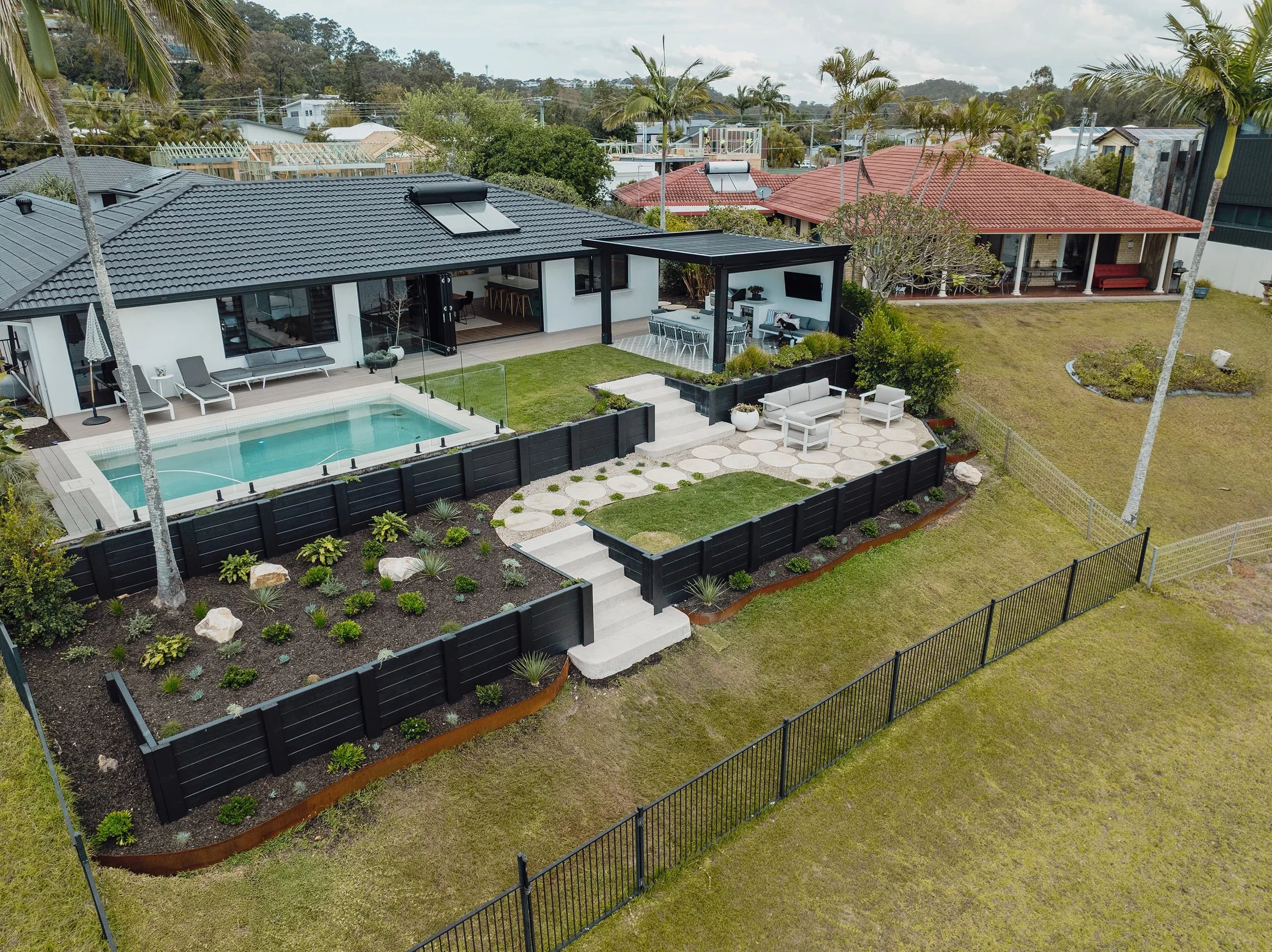 A backyard with a pool enclosed by a black fence, a patio with outdoor furniture, a garden bed with plants, and a house with a gray tile roof and a covered outdoor entertainment area.