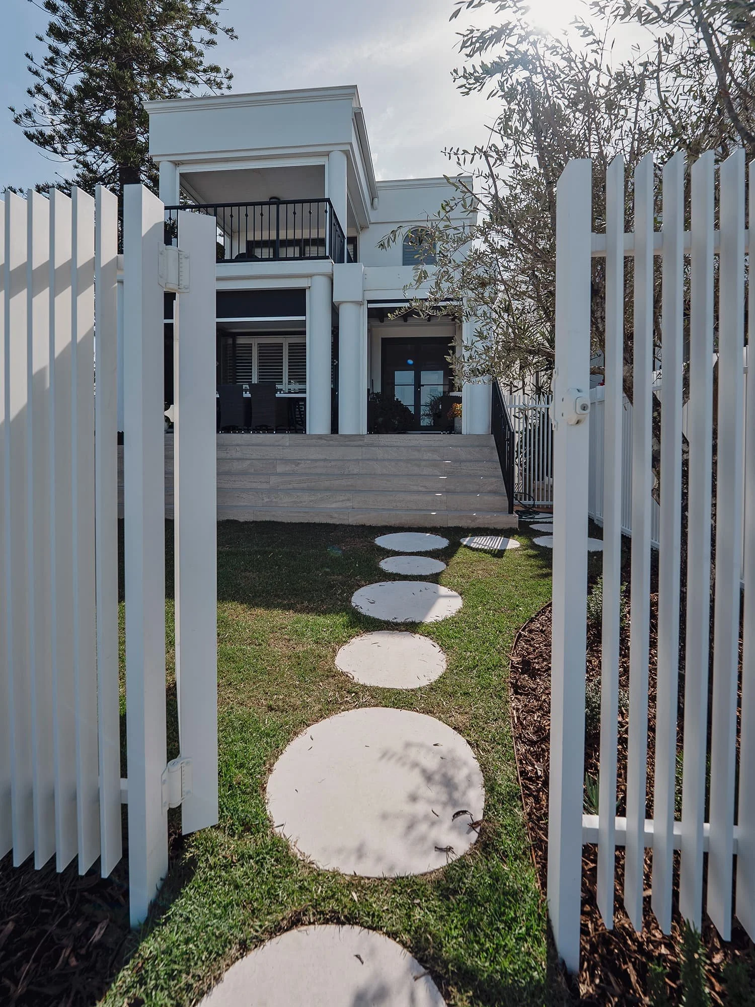View of a modern house with a stone path leading up to a patio. The house has a balcony and outdoor dining furniture. A white gate and a stone pathway lead to the house entrance, surrounded by grass and trees.