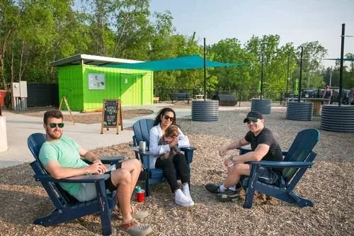 Three people sitting on blue outdoor chairs in a park with a green building and trees in the background.