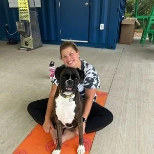 A woman smiling and sitting cross-legged on an orange mat with a big black and white dog in front of her inside a room with blue and green walls.