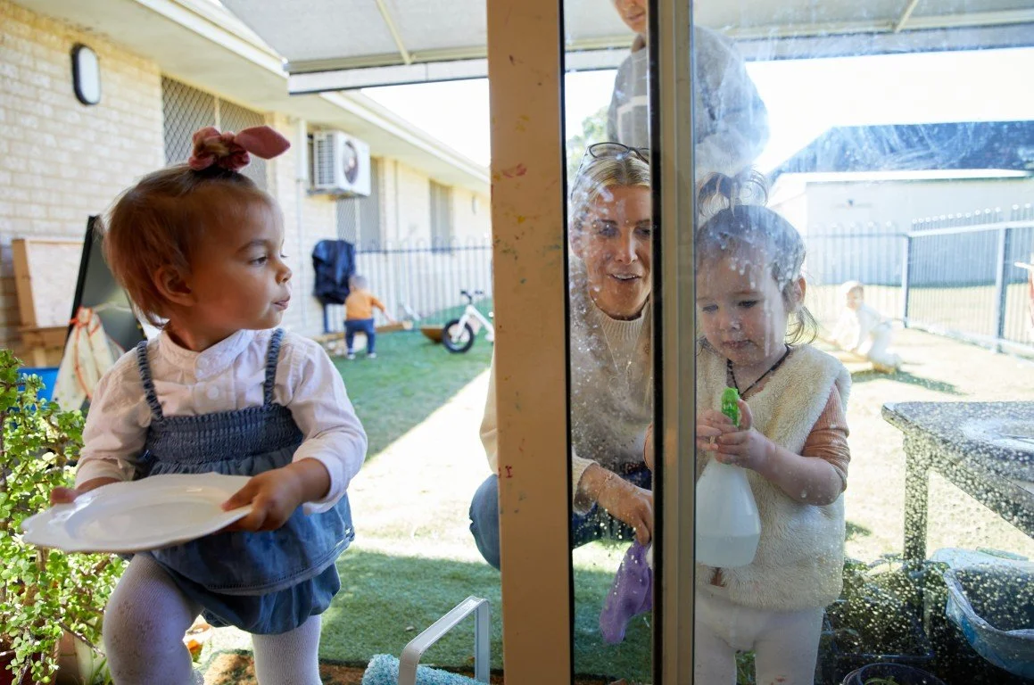 A toddler at our playgroup is putting a ceramic plate away while another is cleaning the glass window