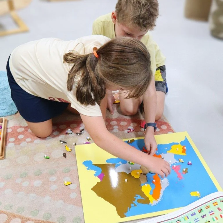 Two primary school aged children, a girl and a boy, are kneeling on a polka-dotted rug, working on a large world map puzzle.