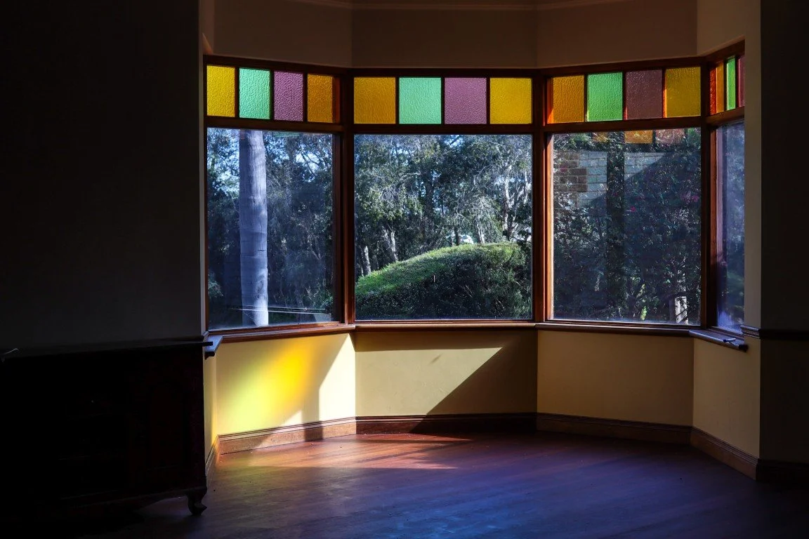 A room with a large bay window featuring colored glass panels at the top, showing outdoor trees and bushes, with sunlight casting a shadow on the yellow walls and wooden floor.